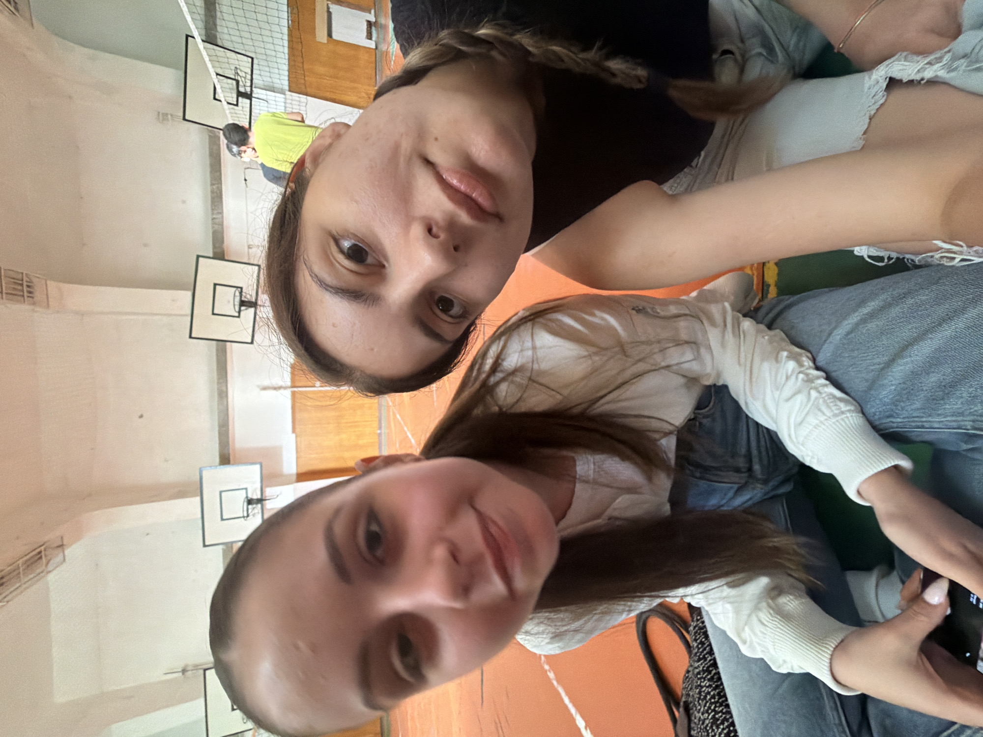 This rotated image features two young women posing for a casual photo, likely a selfie, within an indoor sports hall in Burgas, Bulgaria.
The woman positioned towards the top of the frame has dark brown hair, with a visible braid, and is wearing a black top. She looks directly at the camera with a neutral to slightly smiling expression. The woman lower in the frame has long dark brown hair and a clear smile, also looking at the camera. She is dressed in a white, ribbed long-sleeved top and blue jeans, and her right hand, adorned with light-colored manicured nails, is holding a black smartphone.
The background indicates an indoor gymnasium or sports facility. The floor is a distinct orange-brown color, and the walls are light beige. Multiple white basketball backboards with black square outlines and hoops are mounted on the walls. A white net, likely for sports like volleyball or badminton, is also visible in the distance. Another individual wearing a lime green shirt is partially seen in the background, facing away from the camera.
The photo conveys a relaxed and friendly atmosphere. The indoor setting and artificial lighting do not reveal the time of day or external weather conditions. No discernible text is visible on any objects or clothing within the image.