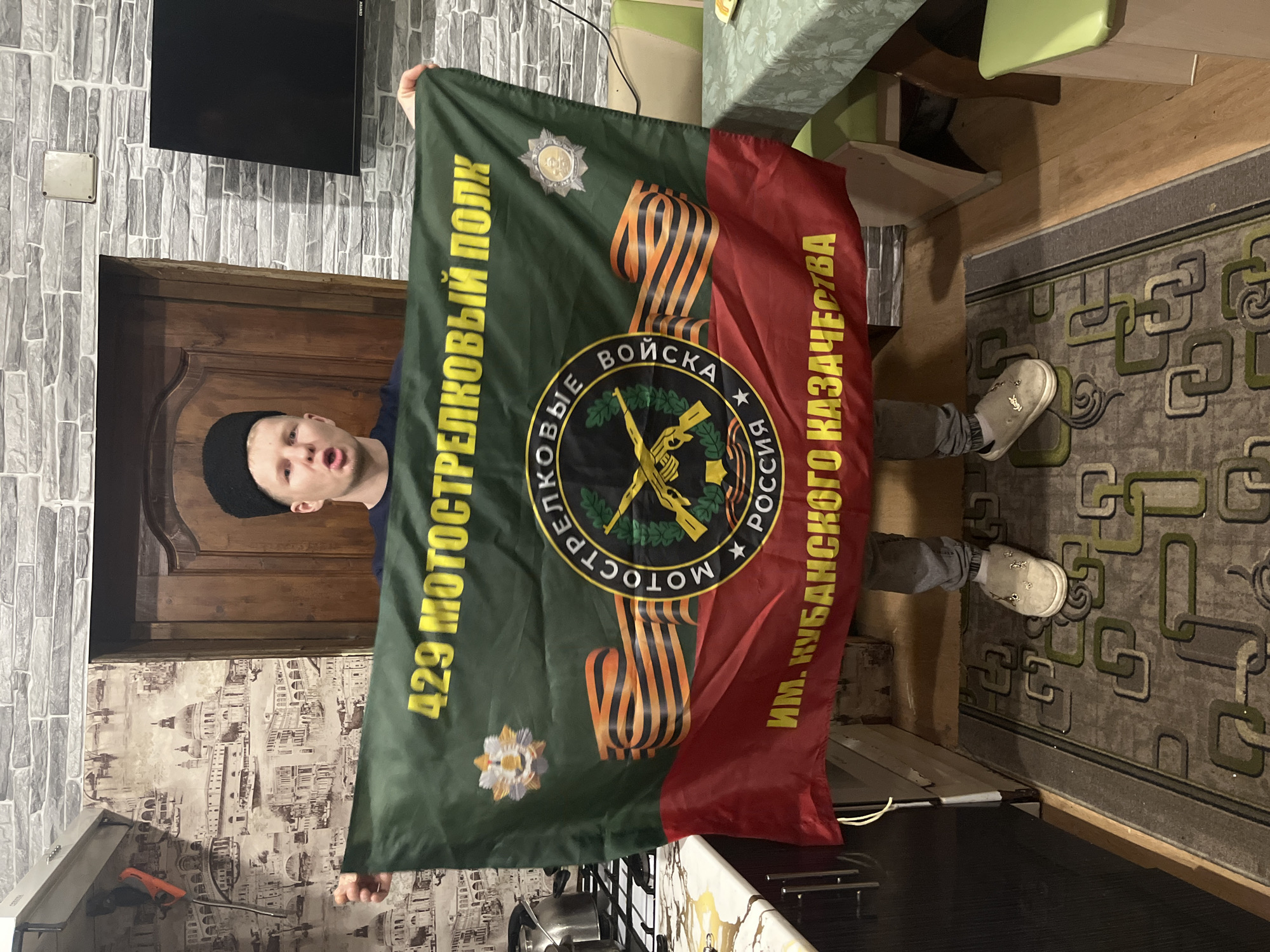 A man wearing a black beanie is holding a flag in front of him. The flag is split in color, with a green section on the left and a red section on the right. On the green section, there is yellow text that reads "429 МОТОСТРЕЛКОВЫЙ ПОЛК" vertically, and a badge at the top left, and a floral emblem at the bottom left. There is an orange and black ribbon draping from the top to the middle. In the center of the flag, there is a black circle containing a rifle. Within the circle, text in Russian reads "МОТОСТРЕЛКОВЫЕ ВОЙСКА РОССИЯ". On the red section of the flag, yellow text vertically spells out "ИМ. КУБАНСКОГО КАЗАЧЕСТВА". The man is standing in an interior space with a door frame and a brick-patterned wall behind him. The floor is carpeted with a geometric pattern. The man is wearing grey pants and light-colored shoes. The location cannot be verified based on the given image alone. Therefore, the city of Vesyoloye, Russia, cannot be confirmed.
