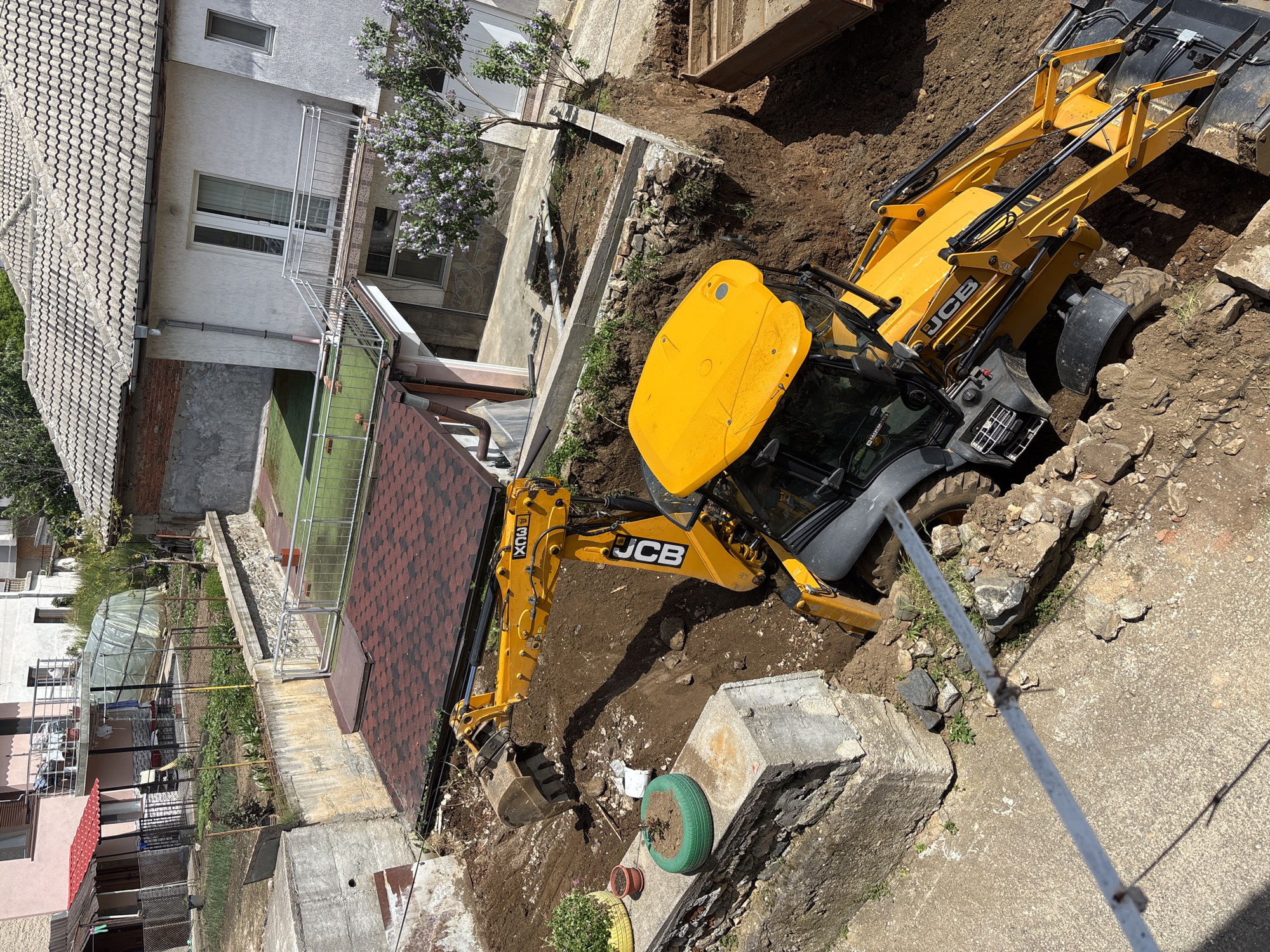 The image shows a yellow JCB backhoe excavator working in a construction zone. The location appears to be Zlatograd, Bulgaria based on architectural style. The backhoe is partially situated in a trench, with its digging arm extended towards a small garden area containing potted plants and a green tire planter. To the left of the garden area is a small building and an area enclosed with safety fences. Other buildings are visible in the background. The environment consists of concrete, dirt, and foliage. There is no visible indication of activity and no presence of people.