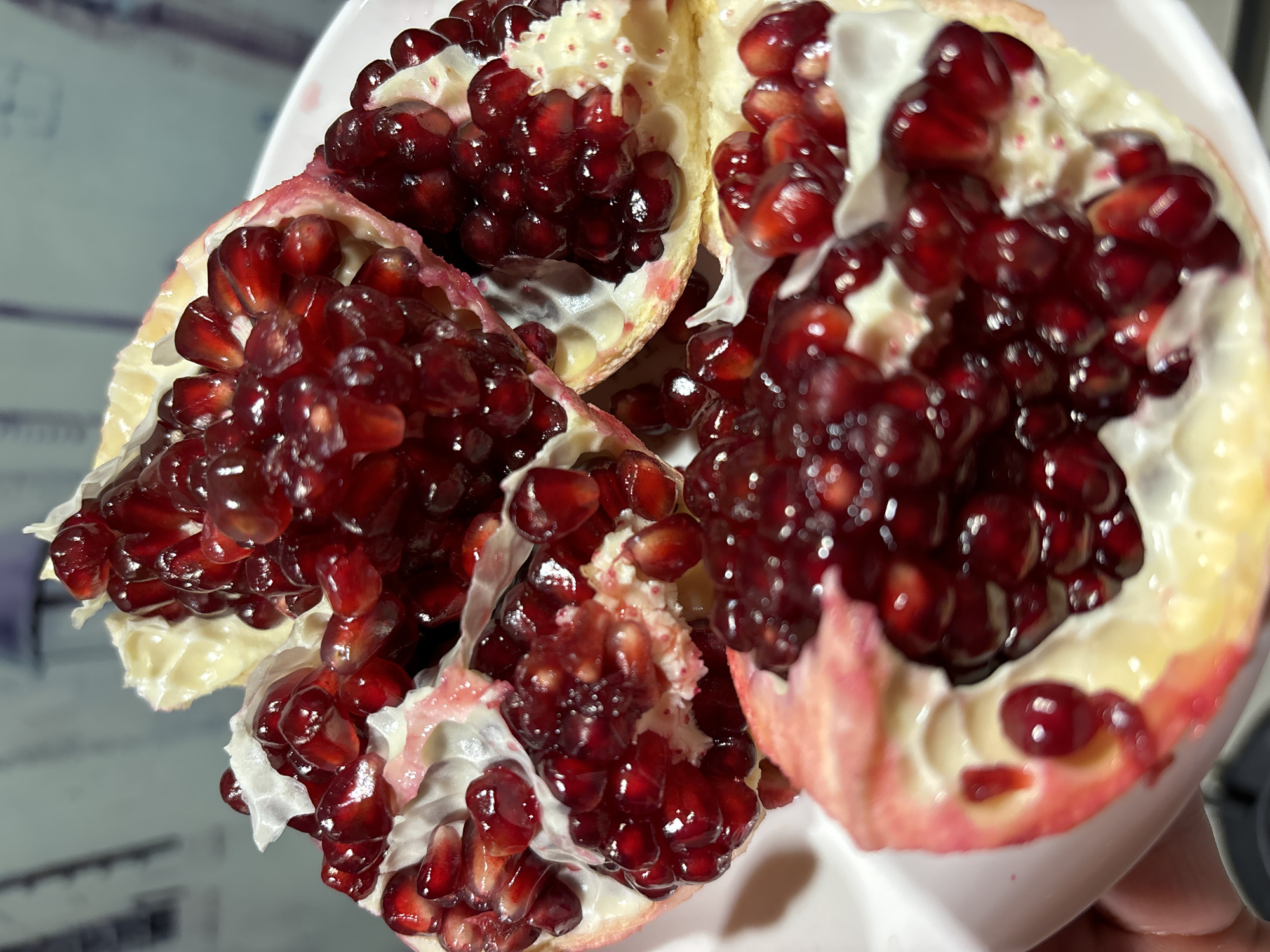 A close-up photograph shows a partially opened pomegranate fruit in a white bowl. The pomegranate is divided into sections, exposing clusters of red arils. The fruit's outer skin is light yellow to pink, with internal white membranes separating the sections. The scene's location in Chaikovsky, Russia, is not discernible from the image.
