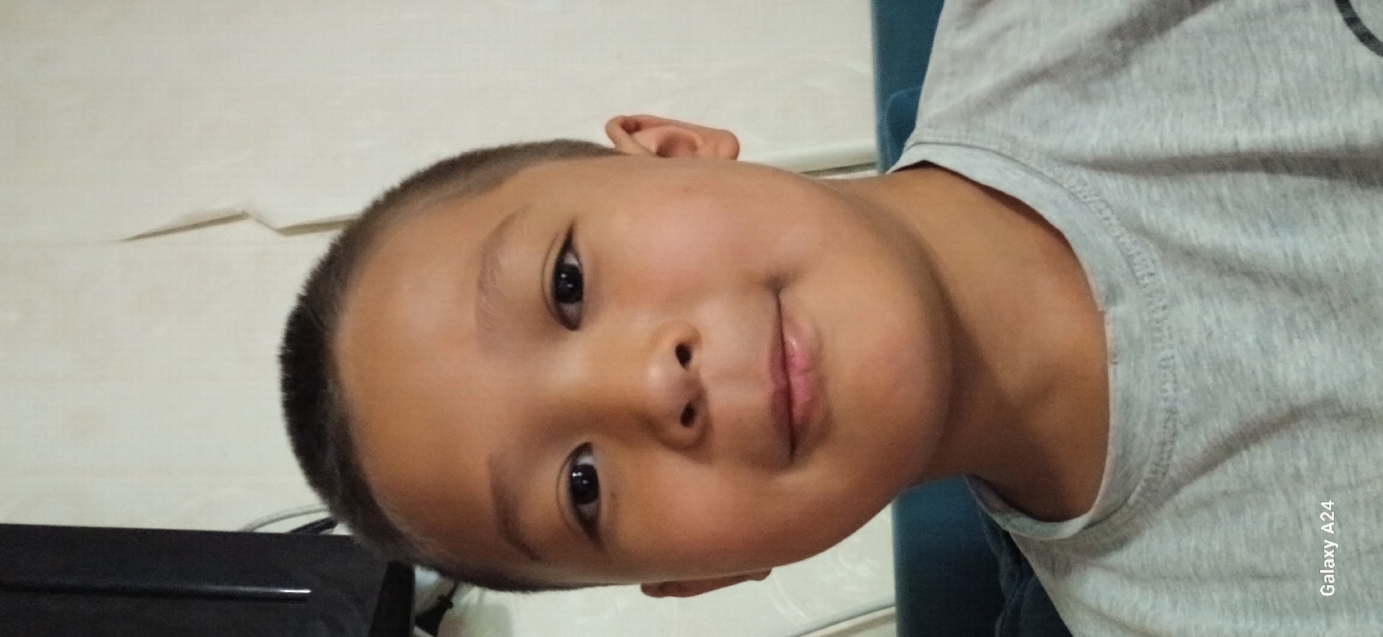A close-up, head-and-shoulders selfie-style shot features a young boy with a short buzz cut and light brown skin. He is looking directly at the camera with a neutral expression, his eyes are dark brown, and his lips are slightly parted. He is wearing a light gray t-shirt. The background is an off-white, textured wall, with a dark vertical object, possibly a television, visible on the far left of the frame. The bottom left corner of the image displays the text "Galaxy A24". The lighting is even, suggesting an indoor setting. The scene is devoid of any specific identifiable landmarks or clues that would pinpoint a location within Bishkek City, Kyrgyzstan.