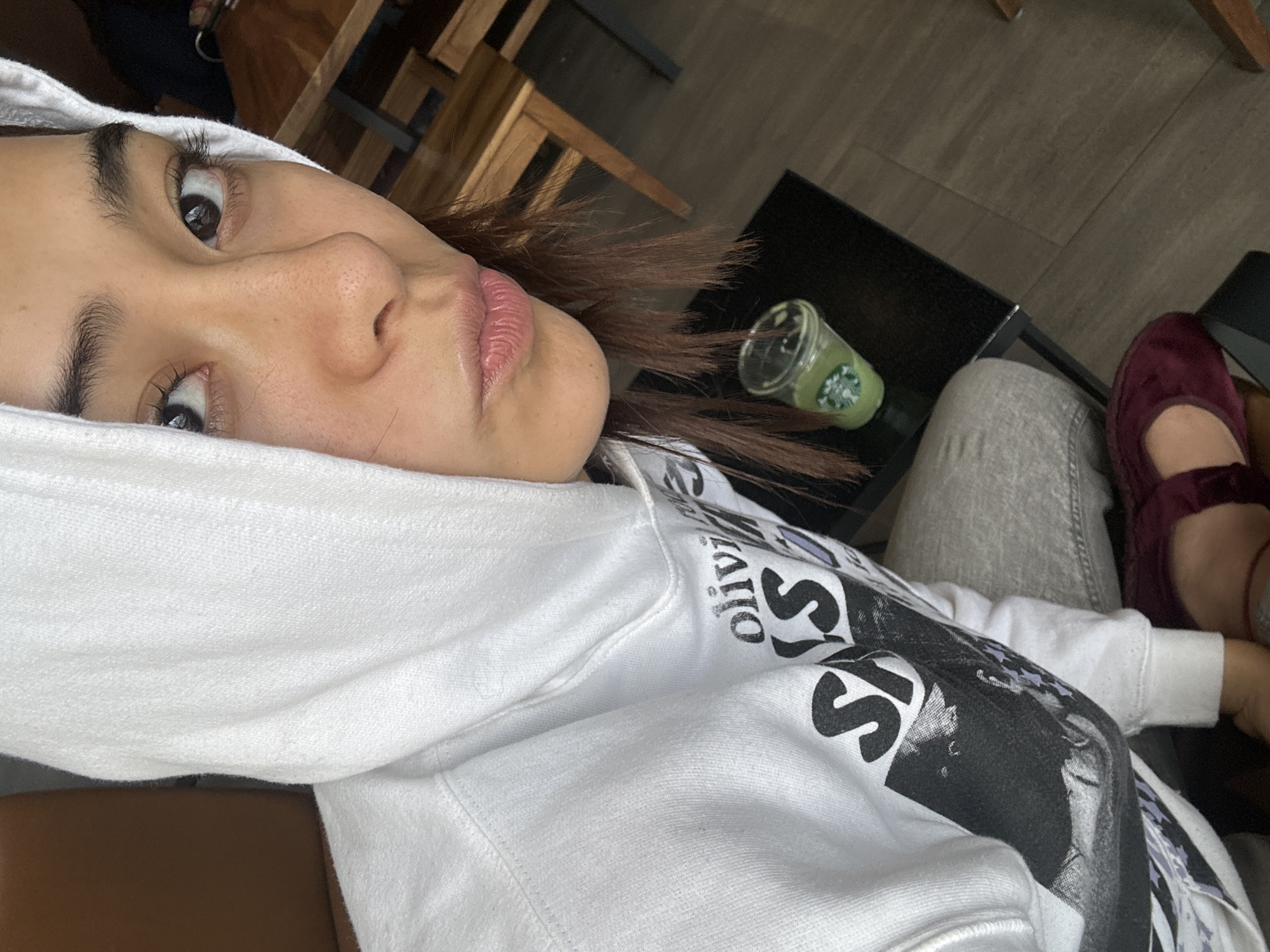 A person wearing a white hooded sweatshirt is visible in a selfie-style shot, possibly in a cafe. A Starbucks cup and a pair of maroon slippers are visible on a small table near the person. The environment appears to be a cafe, based on the presence of furniture and flooring. The exact location in Puebla, Mexico, is not discernible from the image alone. No specific actions or interactions are depicted.