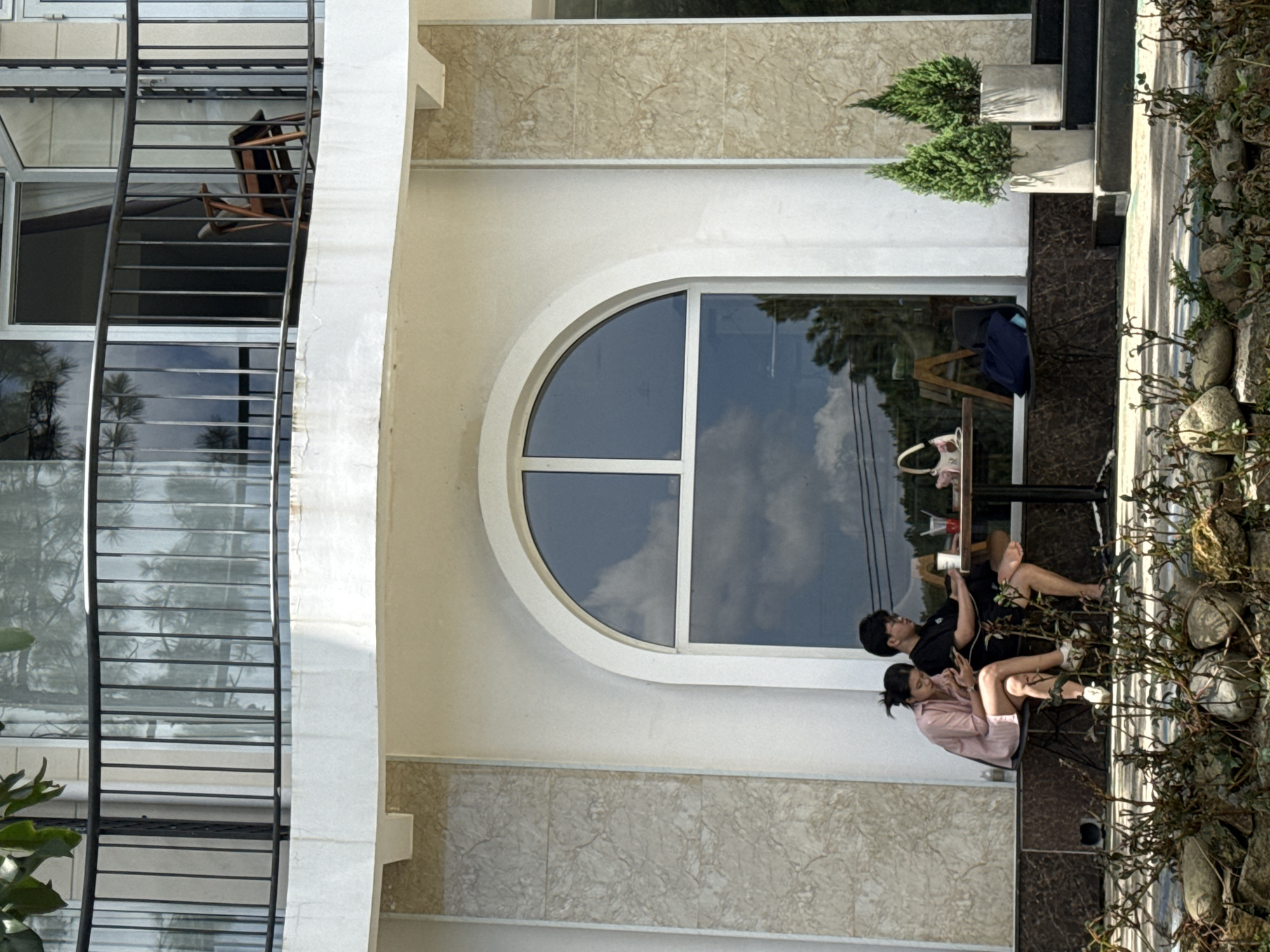 In Da Lat, Vietnam, two individuals are seated at a table outside a building with an arched window. A woman on the left, wearing a pink shirt, is looking at her phone. A man on the right, wearing a black shirt, is also looking at his phone. They are sitting on chairs at a table with drinks and a bag on it. In the background, there are two potted evergreen trees. The building has a balcony with a railing on the upper level. The foreground shows rocks and plants.