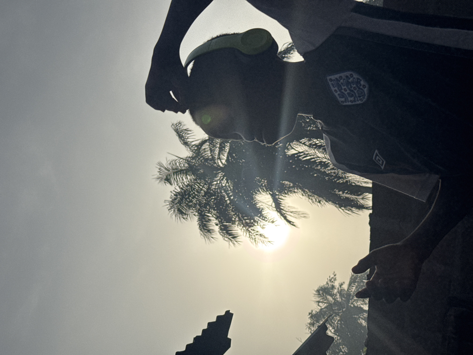 A photograph captures a silhouetted individual, seen from the mid-torso up, positioned against a bright, hazy sky. The person is oriented towards the right, wearing light green/yellow headphones and a dark-colored sports jersey. On the left chest of the jersey, the white three-lions crest of the England national football team is visible. A faint, square logo is also present below the crest. One arm is raised, with the hand near the person's head, while another hand is partially visible further down on the right.
The scene is intensely backlit by the sun, which appears as a strong glare and light streaks in the center-right of the frame. Several palm trees are silhouetted against the brilliant sky behind the individual, their fronds distinctly visible. In the lower parts of the image, dark, angular structures, also in silhouette, suggest architectural elements or ruins.
The lighting indicates it is daytime, with the sun positioned to create a dramatic silhouette effect. The sky appears clear but slightly hazy due to the sun's intensity. The presence of palm trees aligns with the tropical climate of Eziudo, Nigeria, providing a strong geographical cue. The image emphasizes form and contrast, evoking a sense of activity or contemplation under the bright sun.