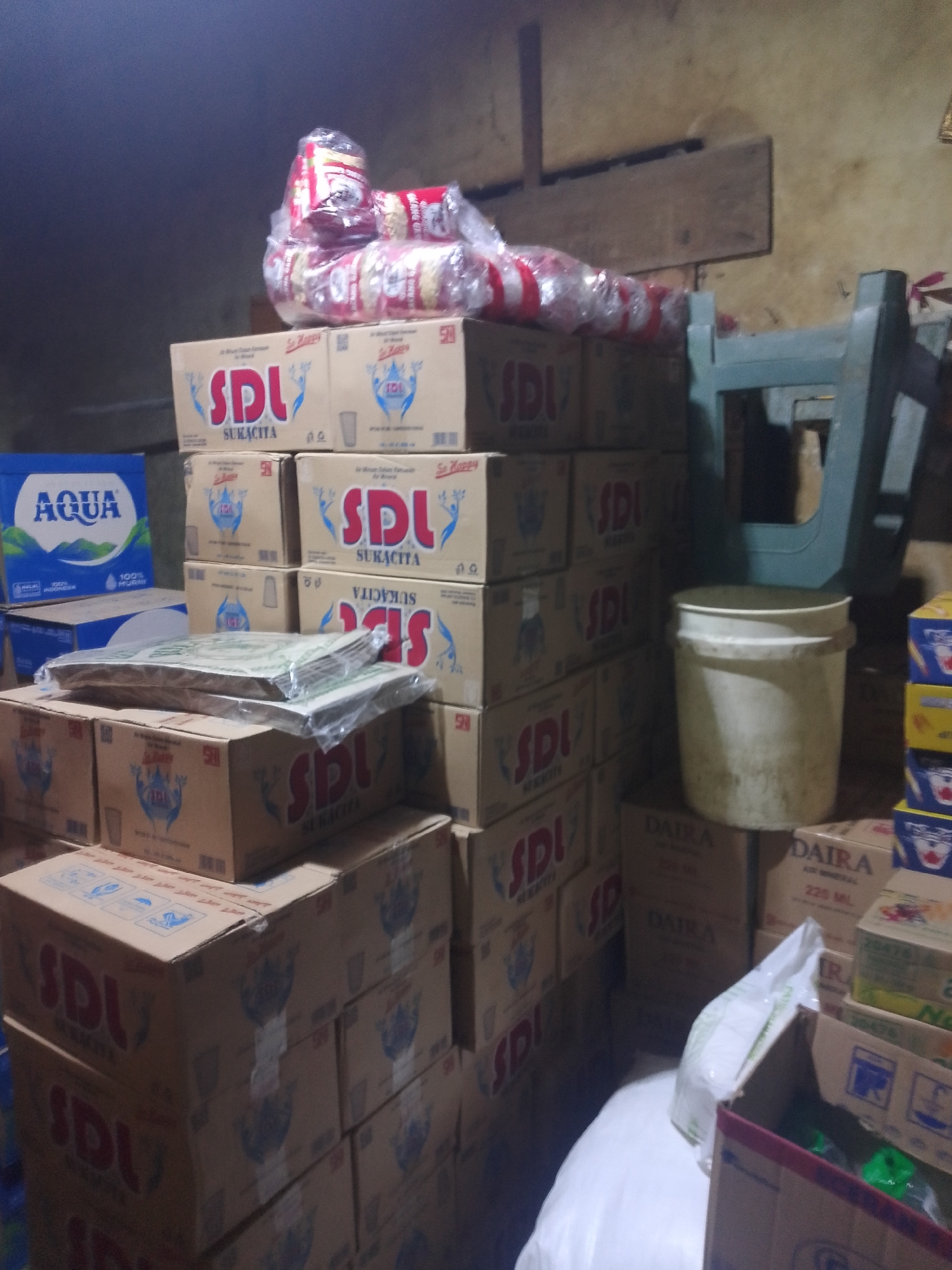 The image depicts an indoor storage area, possibly a warehouse or a stockroom, filled with stacked boxes and various items. In the foreground, several cardboard boxes are piled high, many bearing the prominent "SDL SUKACITA" branding. To the left, a blue cardboard box with the "AQUA" logo is visible, suggesting bottled water. There are also stacks of what appear to be plastic-wrapped goods, possibly snacks or other food items, resting on top of some boxes. In the background, a pale green plastic chair and a dirty white bucket are present. The walls appear to be rough concrete or plaster, and wooden planks are affixed to the wall. The lighting is dim, creating shadows and giving the scene a somewhat cluttered and utilitarian feel. The presence of "AQUA" and the general appearance of the stacked goods suggests a retail or distribution environment. Without further context or identifiable landmarks, it is impossible to pinpoint the specific location in Palembang, Indonesia.