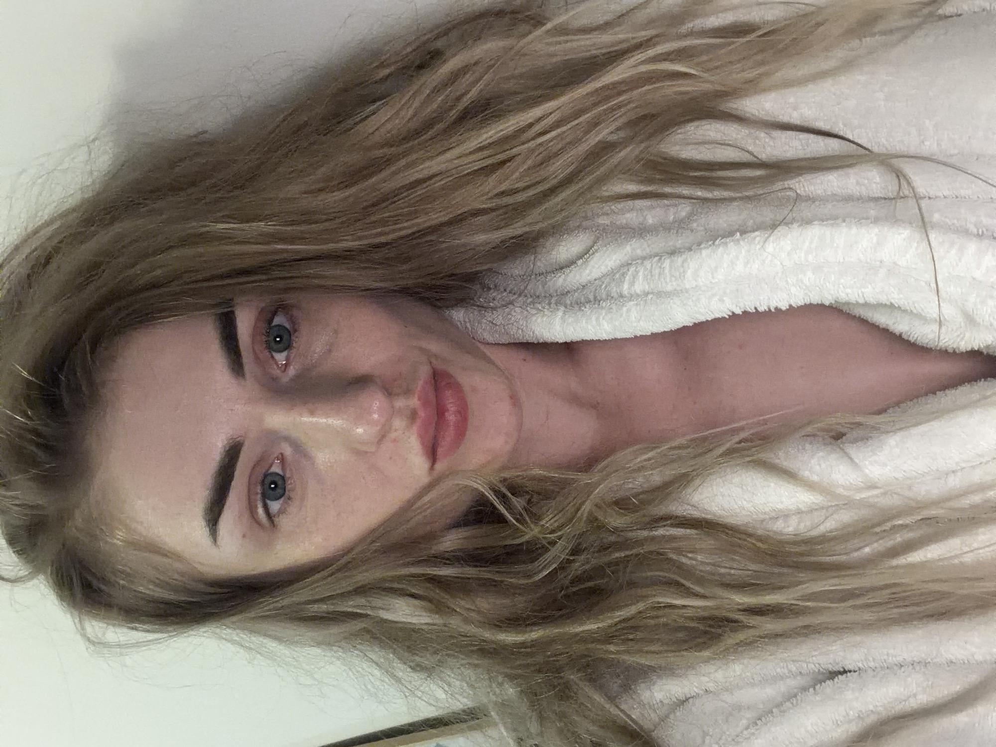The image is a close-up, selfie-style photograph of a young Caucasian woman with long, wavy blonde hair. She is wearing a white, fluffy bathrobe. Her eyes are open and looking towards the camera. Her lips are slightly parted, and she has light makeup on. The background appears to be a plain, light-colored wall. There are no discernible landmarks or environmental features that would indicate a specific location in a city.