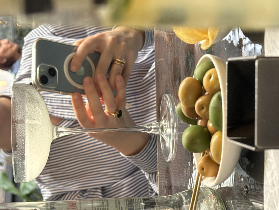 A candid, slightly out-of-focus close-up shot captures a person seated at a dark table in an establishment located in Milan, Italy. The central subject, partially visible from the chin down, wears a blue and white vertically striped long-sleeved shirt. Their left hand holds the stem of a clear, wide-mouthed coupe glass containing a sparkling, pale yellow beverage. Their right hand, adorned with two stacked gold rings on the middle finger and a gold ring with a dark stone on the ring finger, supports a light blue iPhone. The iPhone is encased in a clear cover and features a white circular MagSafe attachment on its back, being held with both hands as if interacting with the device.
On the dark, textured table, a white ceramic bowl holds an assortment of green and yellowish-brown olives, with two small wooden toothpicks visible. To the immediate right, a smaller white bowl containing light-colored snacks is partially visible. A tall, clear glass bottle with an indistinct label stands to the left. A dark rectangular object, possibly a menu or napkin holder, rests in the foreground. In the blurred background, another individual's head and shoulder with reddish-brown hair are faintly discernible, indicating an indoor setting with subtle ambient lighting and hints of greenery.