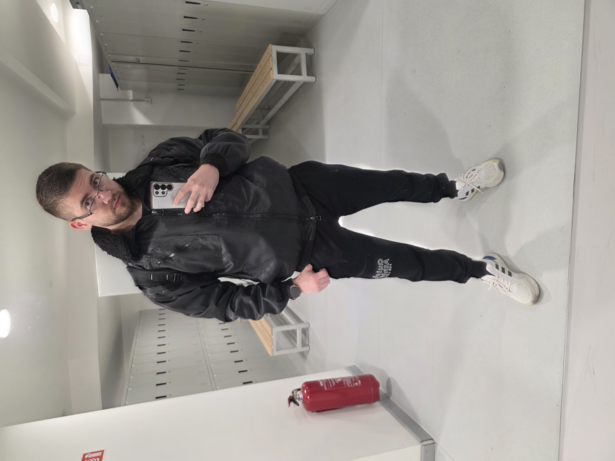 A man is depicted in a mirror selfie, standing in what appears to be an indoor changing room or locker facility. He is positioned centrally, looking directly at the smartphone he holds up with his left hand to take the picture. The man has short dark hair, a beard, and wears glasses. He is dressed in a black zippered jacket with a fur-lined collar, black sweatpants, and white athletic shoes with light yellow and blue accents. A smartwatch is visible on his left wrist. His expression is neutral, with slightly raised eyebrows and wide eyes.
The background, visible in the mirror's reflection, reveals a clean, well-lit interior with light-colored walls and a reflective floor. Rows of grey metal lockers are prominent, and a wooden bench with a metal frame is situated in front of them. Overhead light fixtures are also reflected in the ceiling. In the foreground, a red fire extinguisher is seen lying on a surface near the bottom right of the frame.
White text is partially visible on the man's sweatpants, reading "ONE" and "NBA." The fire extinguisher has unreadable instructional text and symbols. The scene is illuminated by artificial lighting. The location of this facility is Plovdiv, Bulgaria.