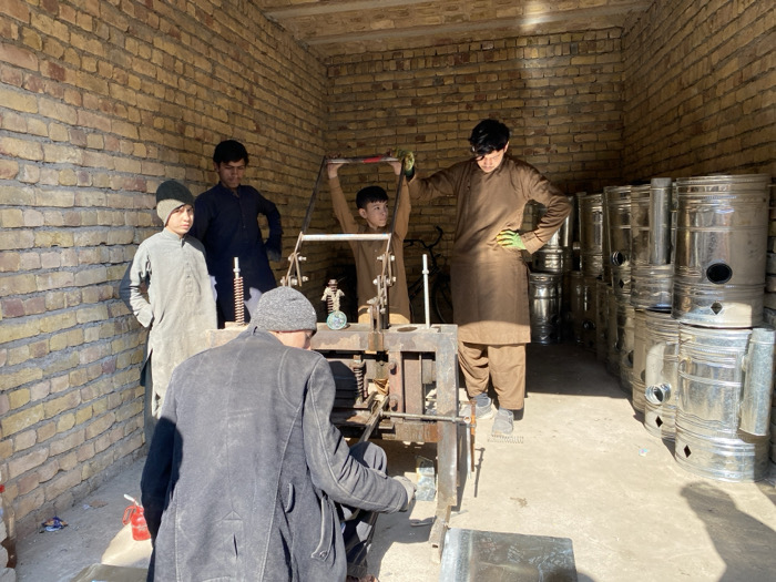 The scene depicts an indoor workshop in Kandahar, Afghanistan, featuring light brown brick walls and a concrete floor illuminated by natural light entering from the right. Five males are present. In the foreground, an adult male, viewed from behind, wears a dark jacket and a grey knit cap while seated or crouched, manipulating a component of a large, manually operated metal press machine. A young male, wearing a light brown tunic, actively operates the machine by pulling down a lever. To his right, an older adolescent or adult male, also in a light brown tunic and green gloves, stands with his left hand on his hip, observing the operation. To the left, two adolescent males stand: one in a light grey tunic and matching knit cap, with hands in pockets, and another in a dark tunic. The right side of the workshop is filled with stacks of cylindrical, galvanized metal stoves or heaters, indicating the manufactured product. A small red object is visible on the floor in the left foreground.