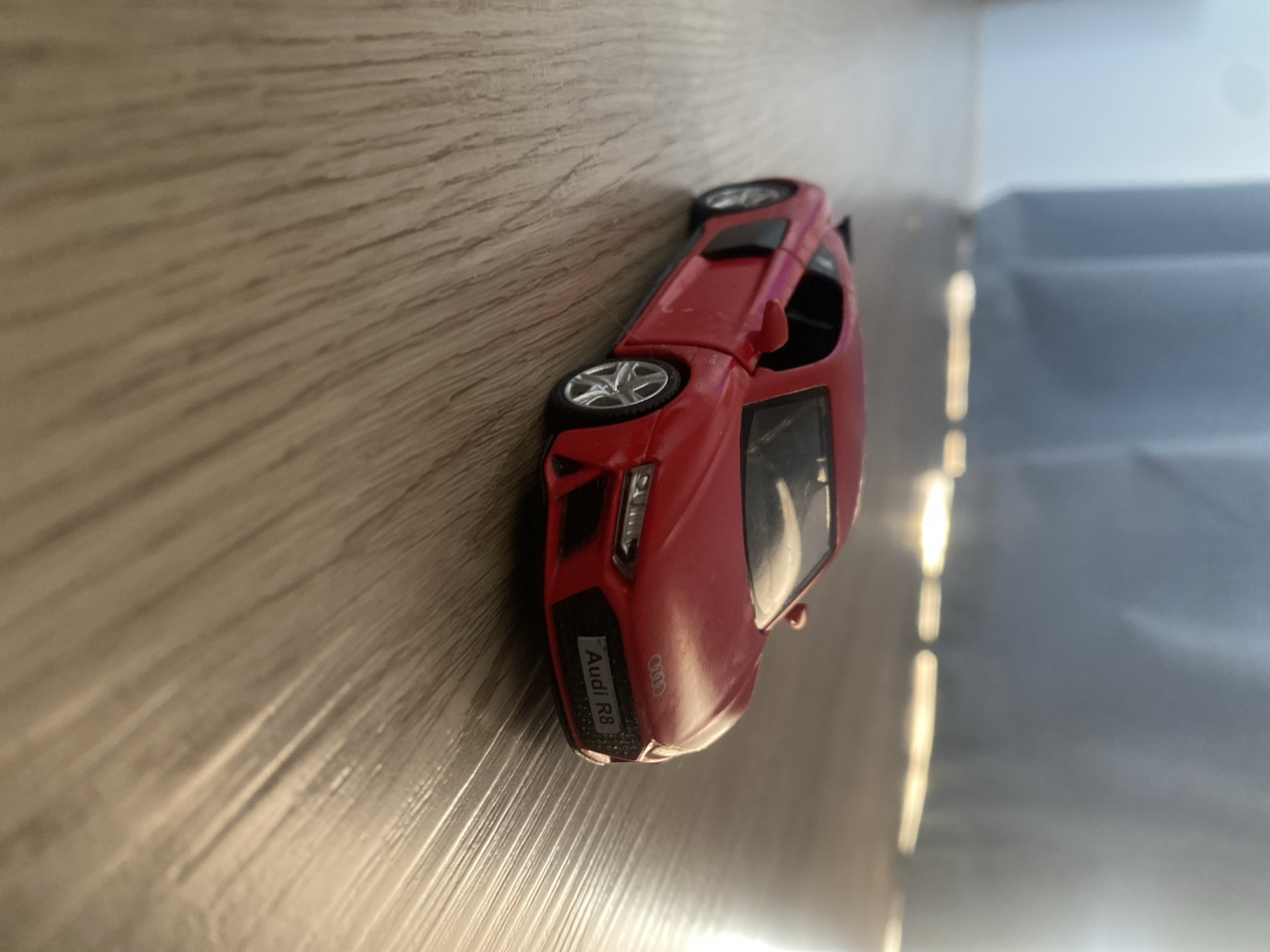 The image shows a close-up, low-angle shot of a red toy Audi R8 car placed on a wooden floor. The car is facing to the right of the frame, with its front slightly angled towards the viewer. The lighting suggests an indoor environment, possibly a room with a window or light source casting a warm glow on the floor and the car. The background is blurred, showing vertical lines that could be curtains or blinds. The floor has a wood grain pattern. There are no people or any other objects visible in the scene. The location in the city of Kozet, Russia, is not identifiable from the visual information provided in the image.