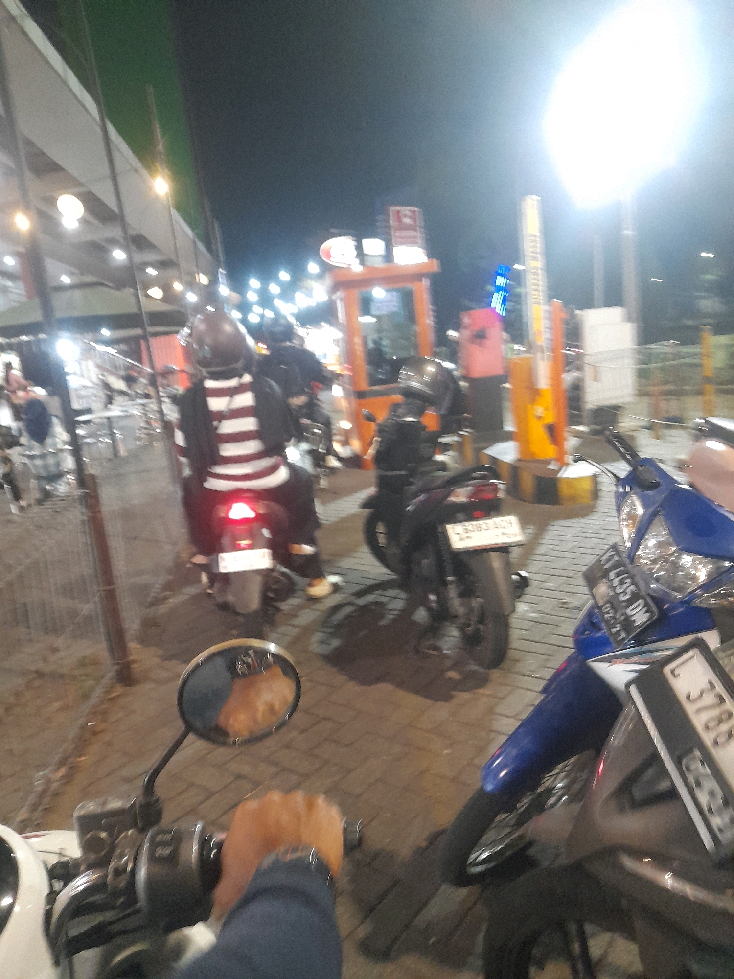 The image shows a night scene with several motorcycles parked or moving on a brick-paved area. In the foreground, a person's hand is visible holding the handlebars of a motorcycle, indicating a first-person perspective. The rider is wearing a dark long-sleeved shirt and a watch. A mirror reflects a portion of the rider's face.
Further back, a person wearing a helmet and a striped shirt is seated on a motorcycle, which has its brake light illuminated. Another motorcycle is parked to the right, with a blue front fairing and a license plate reading "KT 4435 DM". A third motorcycle is positioned behind it, with its license plate partially visible.
The background features a brightly lit structure that appears to be a toll booth or a payment gate, with yellow and orange poles. Lights from various sources illuminate the scene, creating a somewhat blurry and high-contrast effect due to the low light conditions. The presence of multiple motorcycles and the architectural elements suggest this could be a parking area, a public space, or an entrance to a facility in Surabaya, Indonesia, likely a commercial or transportation hub.