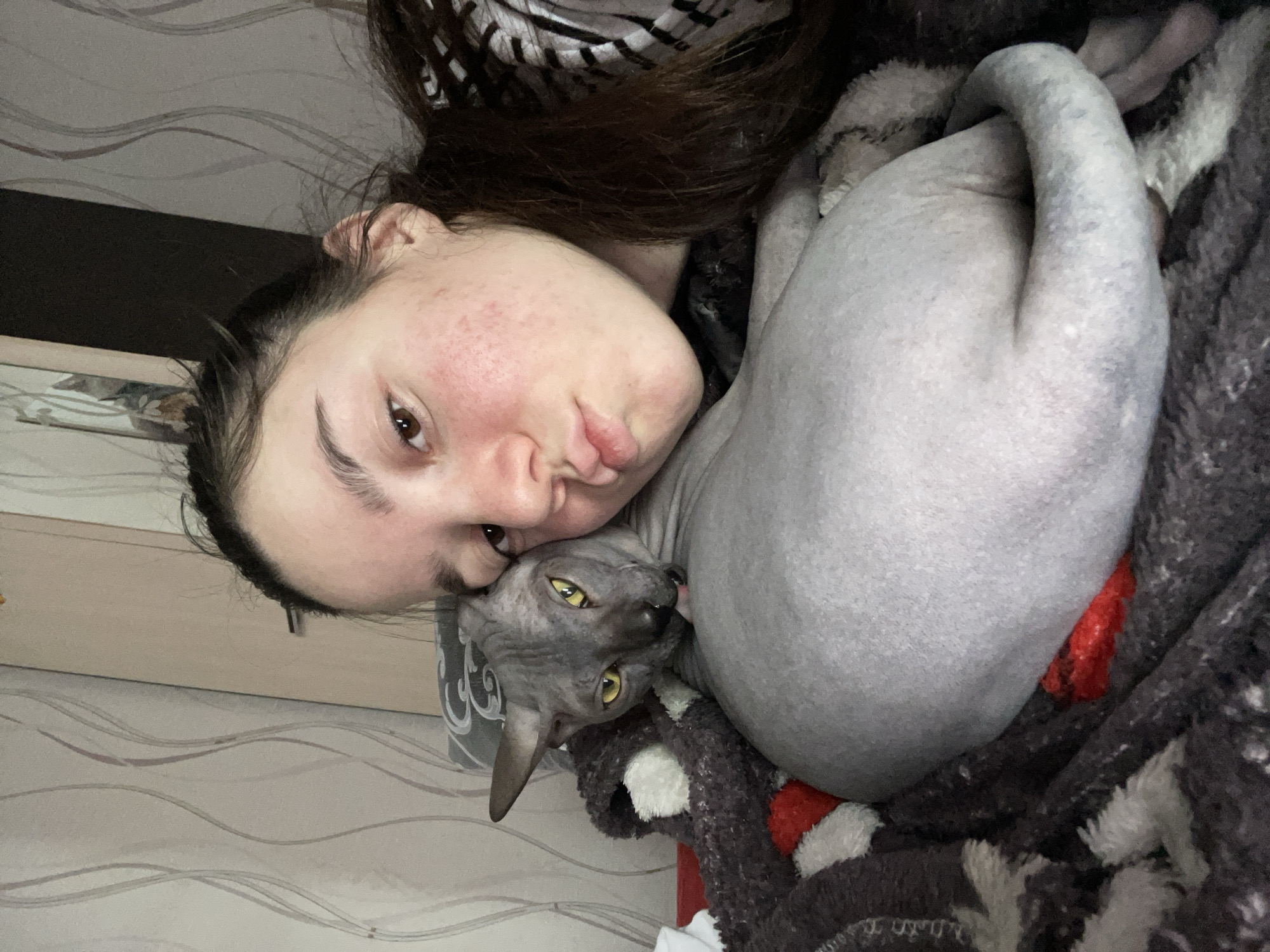 A young woman is lying down next to a gray Sphynx cat. The woman is fair-skinned with brown hair, and she is looking at the camera with slightly pursed lips. Her face is close to the cat's head. The cat is curled up and looking at the camera. The location is likely Ольховка, Russia. They are lying on a bed with patterned bedding and blankets.