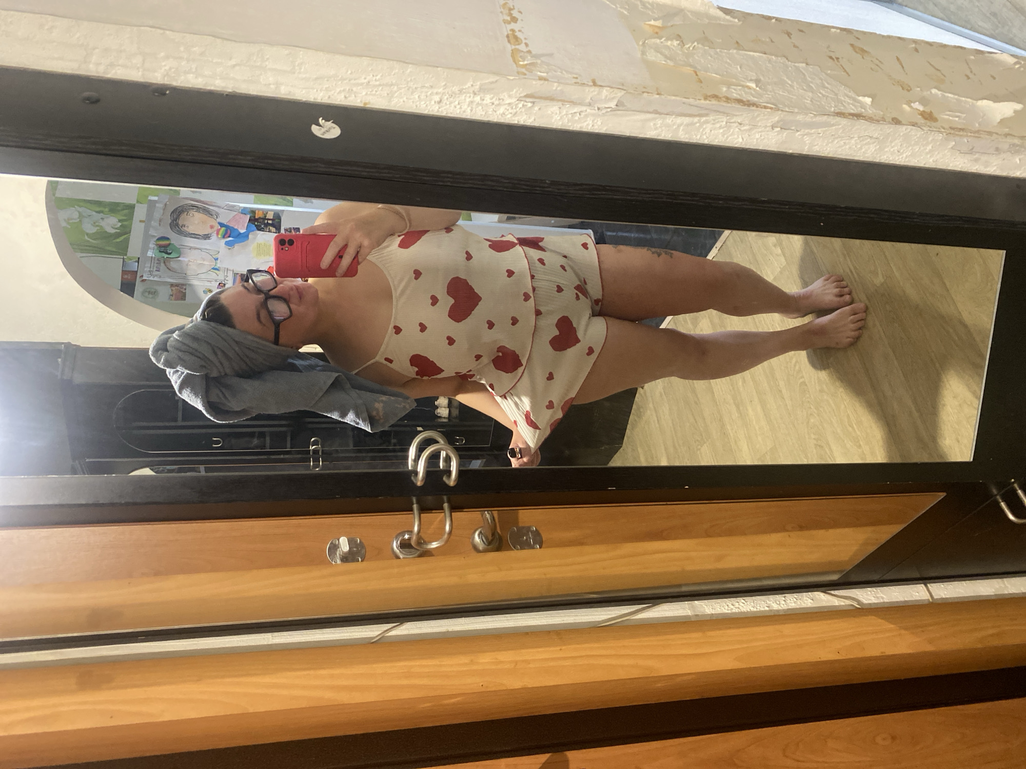 A young woman with a towel wrapped around her head stands in front of a full-length mirror, taking a selfie with her pink smartphone. She is wearing a cream-colored pajama set adorned with small red hearts. The background of the reflection shows a wall covered in children's drawings and a framed picture. To the left of the mirror is a wooden door with silver hardware. The floor is made of light-colored wood. The setting appears to be a residential interior, potentially a bedroom or bathroom, and is not indicative of any specific location within the city of Chaikovsky, Russia.