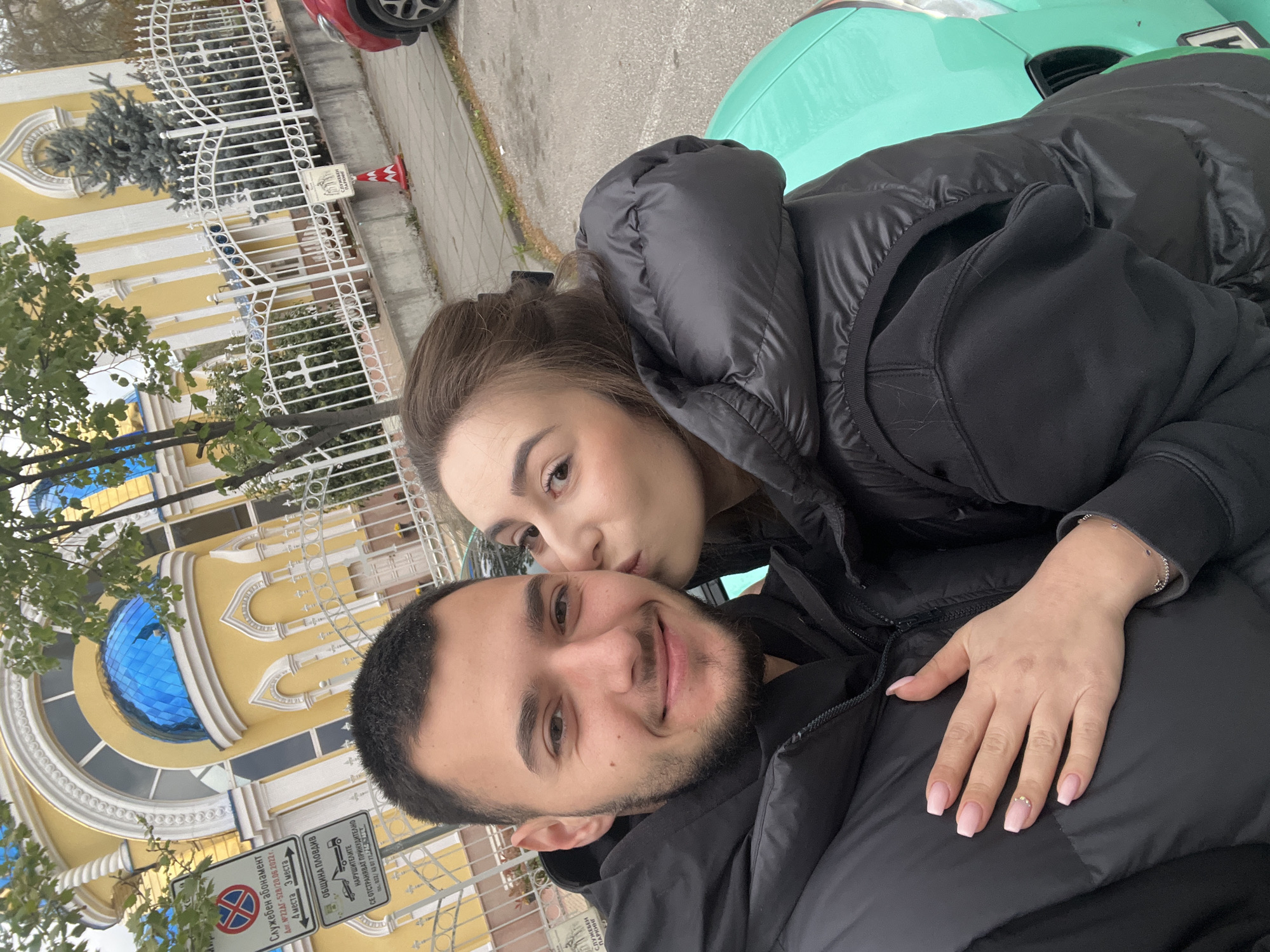 The image captures a tilted selfie of a couple outdoors, likely on an overcast day. In the foreground, a man with a beard and short dark hair smiles gently at the camera, wearing a black puffy jacket. Behind him, a woman with brown hair, also in a black puffy jacket, leans in to kiss his left cheek. Her left hand, adorned with light pink manicured nails, one featuring a small decorative stone, rests on his shoulder. She wears a delicate silver bracelet.
The background features a distinctive yellow building with ornate white architectural elements and a large, blue domed window. A white metal fence with decorative patterns runs along the street, behind which some evergreen trees are visible. Parked cars are partially seen; a bright teal or mint green vehicle is behind the woman, and a red car's wheel is visible at the top. A red and white traffic cone stands near the fence.
Several signs provide textual information and location cues. One prominent sign states "ОБЩИНА ПЛОВДИВ" (Municipality of Plovdiv) and warns, "НАРУШИТЕЛИТЕ СЕ ОТСТРАНЯВАТ ПРИНУДИТЕЛНО" (Violators are removed by force/towing). Another sign designates "СЛУЖЕБЕН ПАРКИНГ" (Official Parking), detailing "Служебен абонамент" (Official subscription) with a contract number (22ΑΓ-528/20.06.2022) and specifying "4 места 3 места" (4 spaces, 3 spaces). A standard "No Parking" sign is also visible. These details confirm the setting as Plovdiv, Bulgaria. The image conveys a tender moment between the couple in an urban environment.