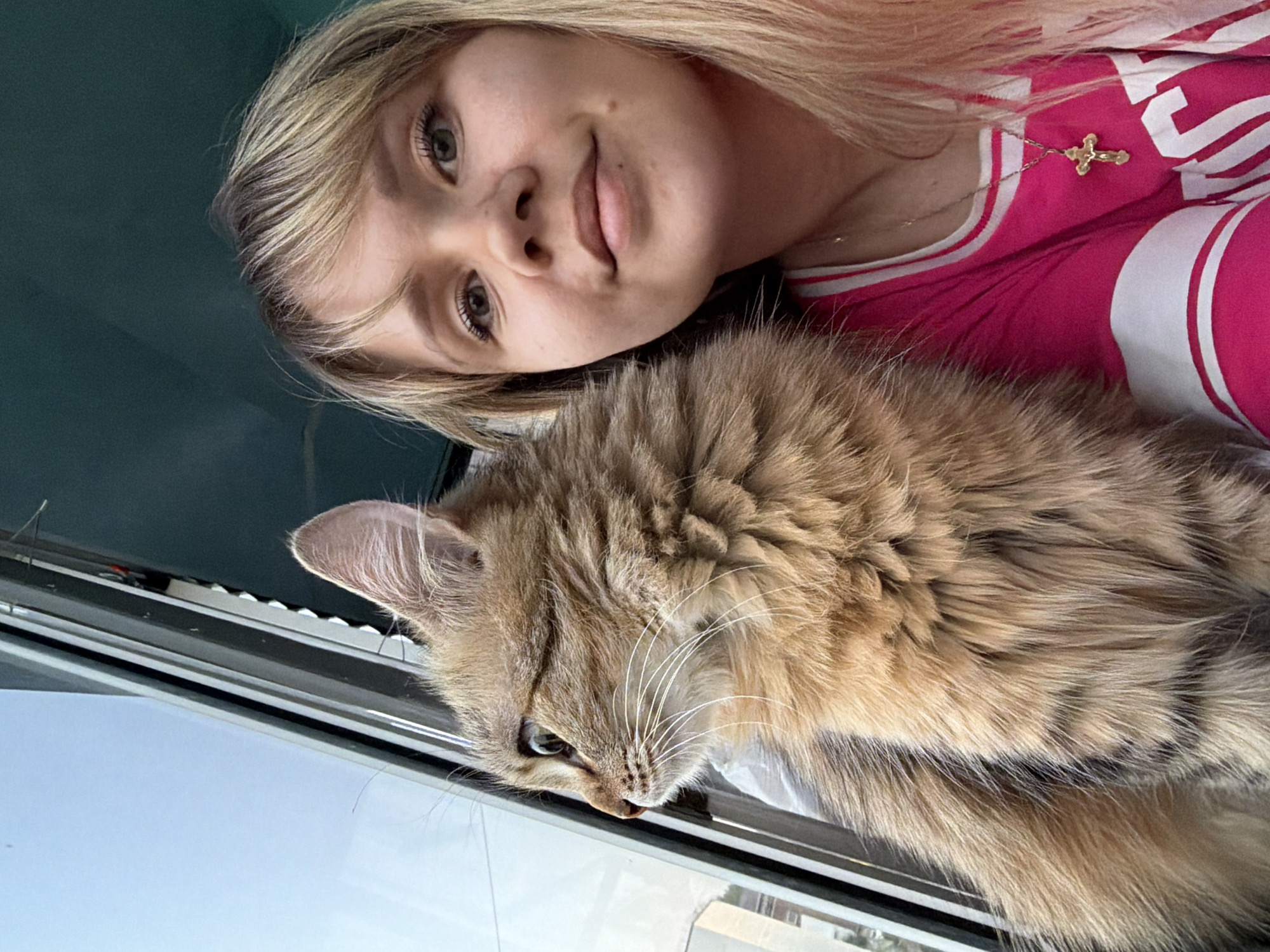 A young woman with blond hair and a pink and white shirt is taking a selfie with a fluffy brown cat next to a window. The location is Chaikovsky, Russia, as evidenced by the visible window frame and external surroundings. The woman is smiling slightly, and the cat is looking down towards the street. A gold cross necklace is visible on the woman's chest. The window appears to be closed.