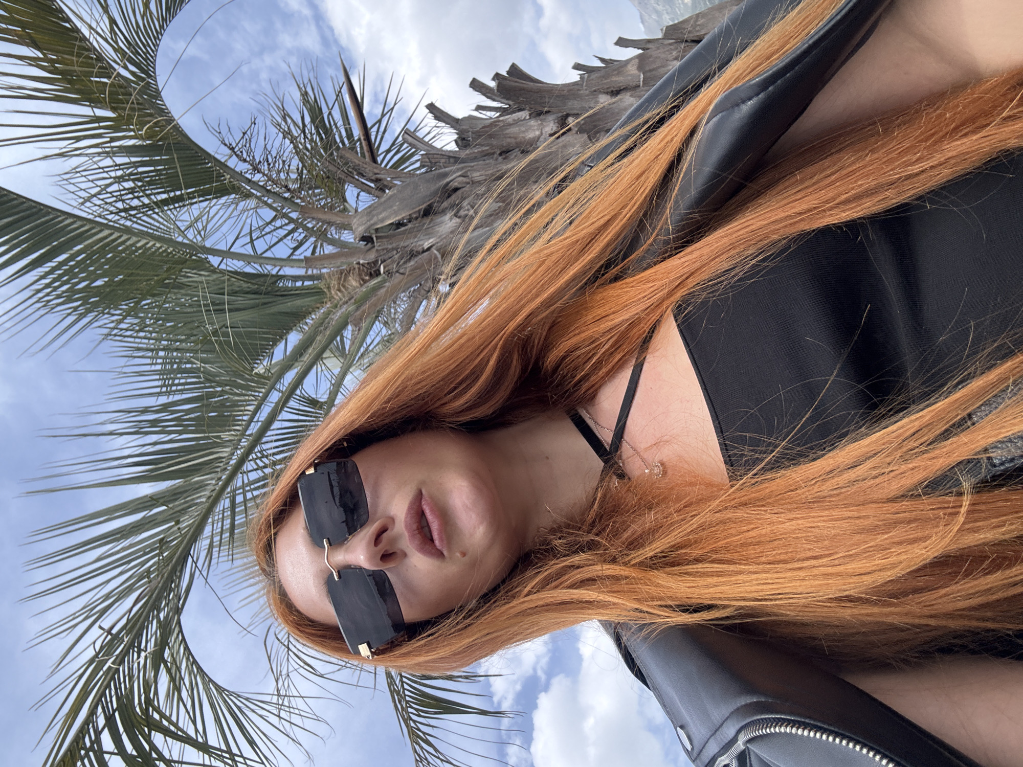 A low-angle outdoor shot features a woman with long, red hair wearing black sunglasses and a black top. She's partially facing the camera, with a palm tree behind her. The sky is visible with scattered clouds. The setting is likely a coastal area due to the presence of the palm tree and sky. The location is in the city of Budva, Montenegro.