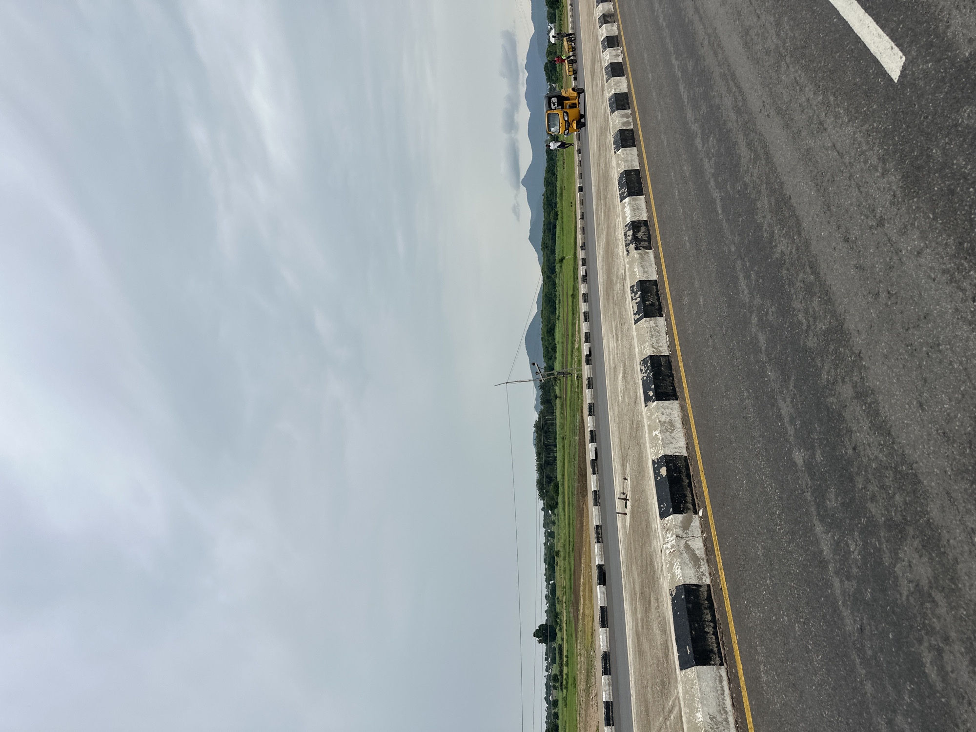 This image depicts a road scene in Jalingo, Nigeria, on a cloudy day. In the foreground, a paved road with a yellow line marking the edge is visible. Adjacent to the road is a concrete barrier with alternating black and white painted sections.
Further down the road, a yellow auto-rickshaw is in motion, carrying several people. To the right of the auto-rickshaw, a group of individuals appears to be pushing carts or moving objects, suggesting some form of transport or labor.
In the background, a vast expanse of green fields stretches towards a range of low, undulating hills or mountains. Sparse trees and vegetation dot the landscape. The sky is overcast with a thick layer of clouds, casting a subdued light over the entire scene. Electrical wires and poles are visible in the middle ground, indicating the presence of infrastructure. The overall impression is one of a rural or semi-rural setting on the outskirts of Jalingo.