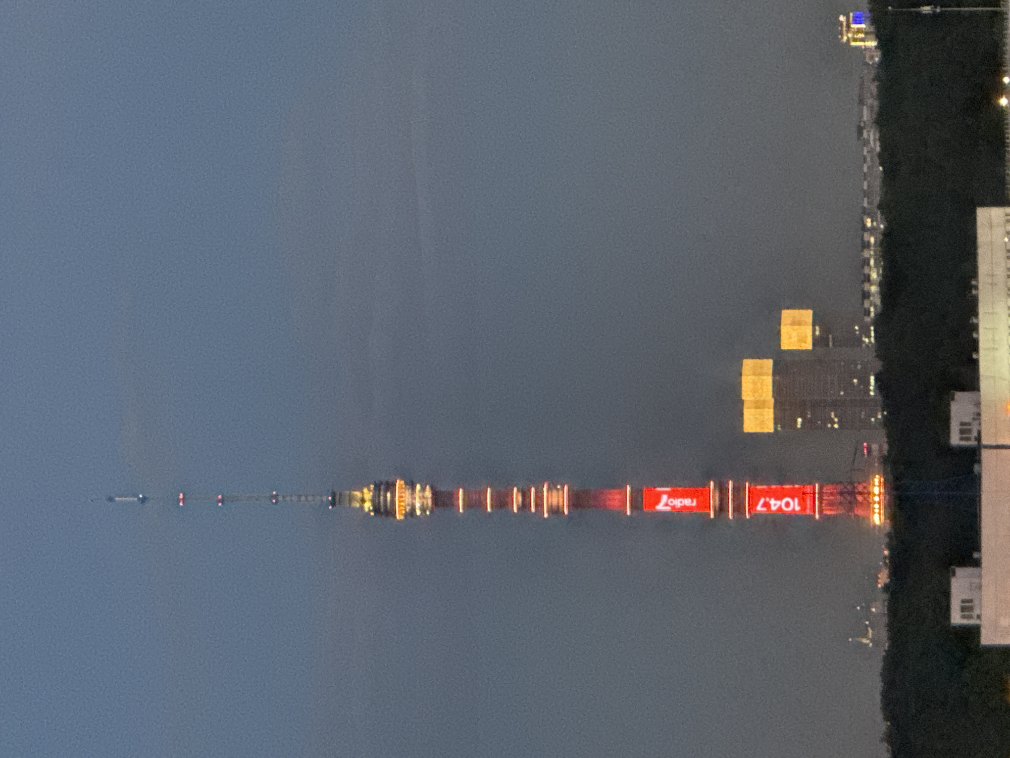 The image shows the Ostankino Tower in Moscow, Russia, at dusk. The tower is illuminated with red and yellow lights. The numbers "104.7" are prominently displayed in red on the tower, along with the word "radio7". In the background, several other buildings and city lights are visible. The sky is a deep blue, indicating the transition from day to night. The scene is framed by a dark line of trees in the foreground, with some white buildings visible at the very bottom of the image.