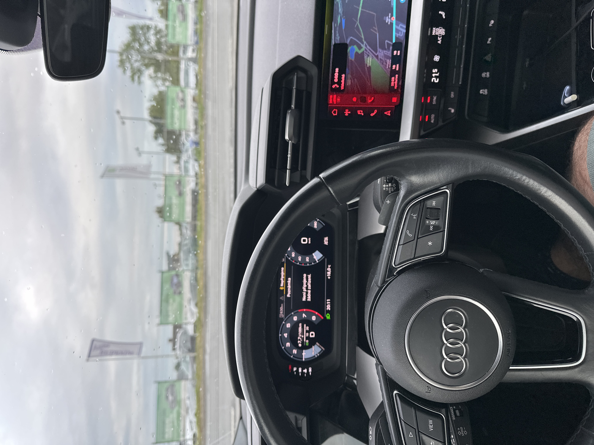 This is a first-person view from inside a car, looking forward through the windshield at a car dealership. The scene is primarily composed of the car's interior: a steering wheel with the Audi logo prominently displayed, a digital dashboard showing speed and navigation information, and a central infotainment screen displaying a map. Various buttons and controls are visible on the steering wheel and dashboard. The digital dashboard shows a speed of 0 km/h, a fuel consumption of 7.7 L/100km, and a time of 20:11. The infotainment screen indicates a navigation route with a destination labeled "Vídeňská" and an estimated arrival time of 20:23. The weather appears overcast and potentially rainy, with water droplets visible on the windshield. Outside the car, rows of vehicles are parked, characteristic of a car dealership lot, with green promotional banners visible in the background. The overall setting is a car dealership on a cloudy day, likely in the early evening, given the time displayed on the dashboard. No people are directly visible in the image.