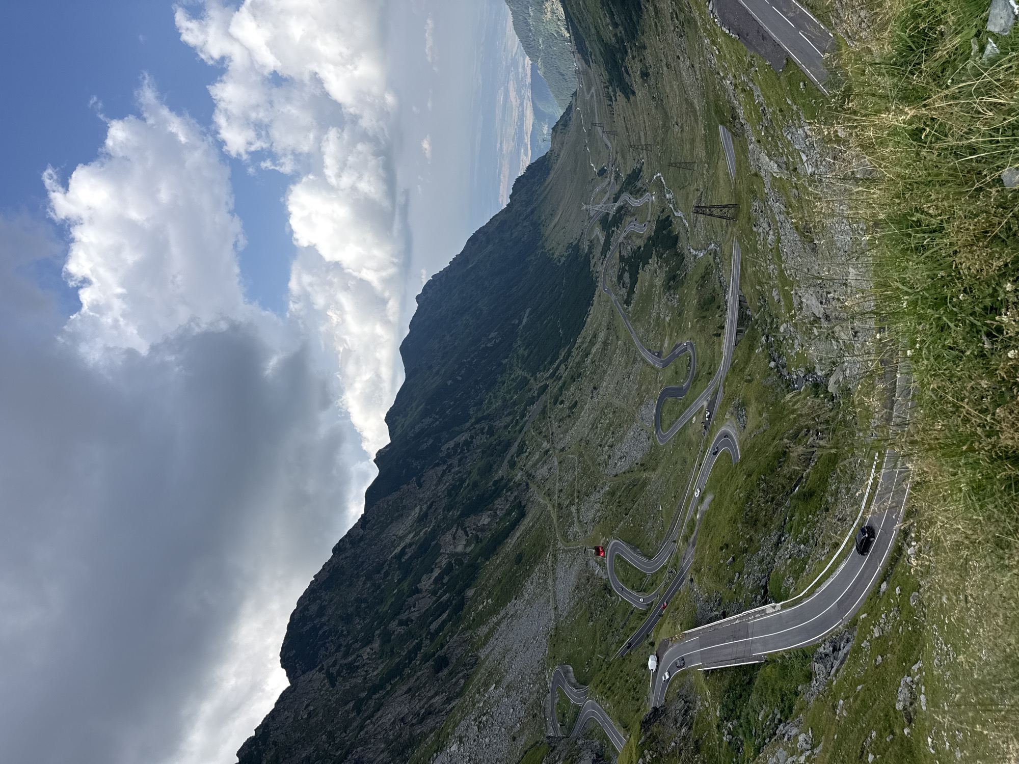 The image displays a dramatic mountain landscape featuring the Transfăgărășan Highway, a winding road in Cârțișoara, Romania. The road snakes through steep, grassy slopes and rocky terrain, with numerous hairpin turns visible. Several vehicles, including cars and a white van, are present on the asphalt road. A prominent red cabin, likely a cable car, is suspended above a section of the road. The surrounding mountains are covered in green vegetation, with patches of exposed rock. The sky is a mix of dramatic clouds and clear blue patches, suggesting variable weather conditions. In the distance, a valley opens up with more mountains and hints of human settlement. The overall scene conveys a sense of rugged natural beauty and the engineering feat of the mountain road.