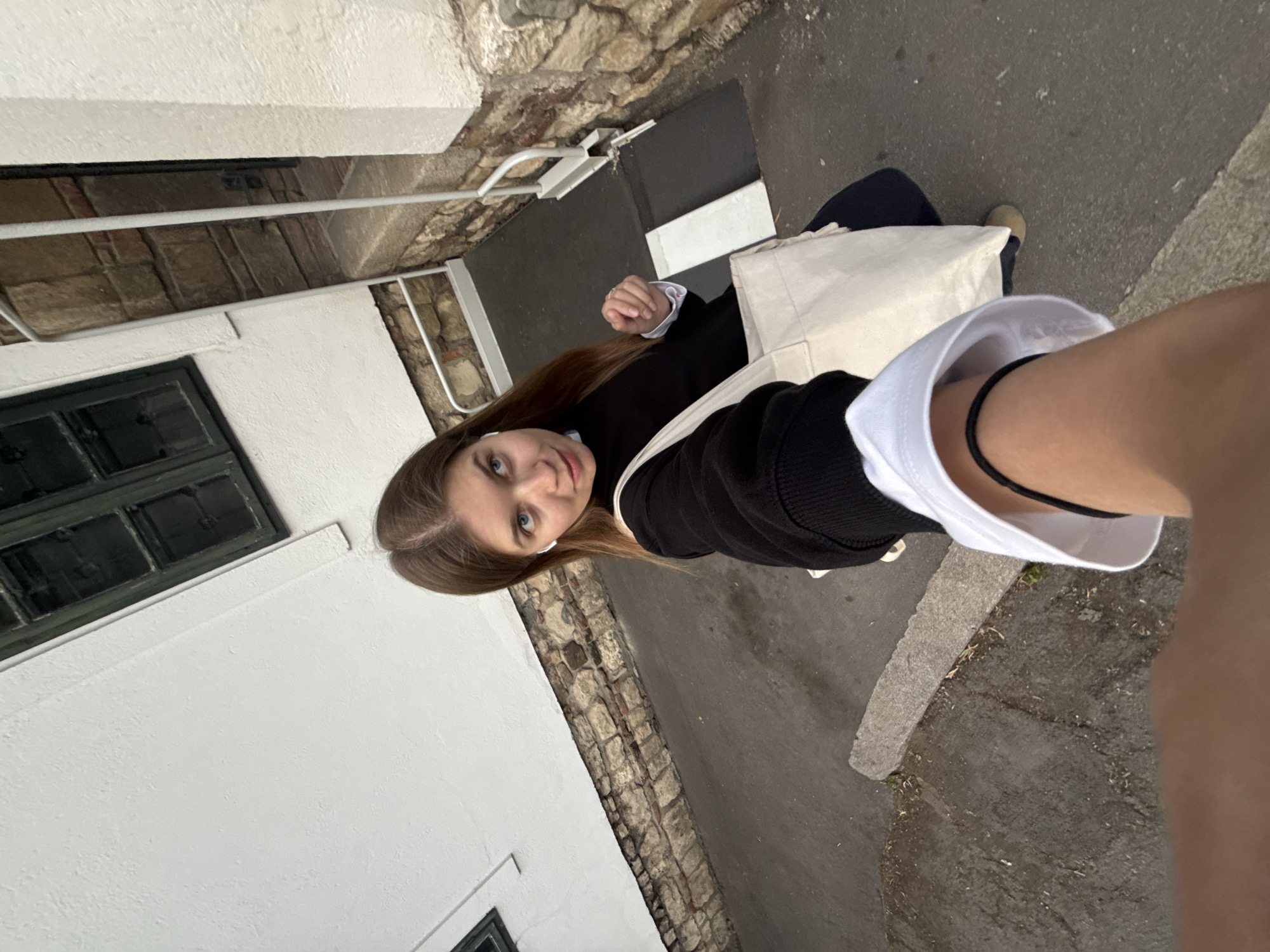 A woman stands in an alleyway. She is wearing a black jacket, and she is carrying a white tote bag. The alleyway is paved with stone, and there is a white building on one side. A stone wall is visible between the building and the alleyway. A window is visible on the building. The woman appears to be taking a selfie. The location is likely in Belgrade, Serbia.