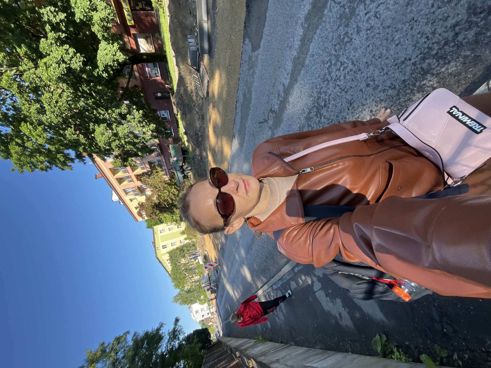 A woman with sunglasses and a brown leather jacket takes a selfie while walking on a sidewalk. A young person in a red hoodie walks ahead of her on the same sidewalk. The street is undergoing construction, with mounds of dirt and construction materials visible on the right side of the road. Buildings line the street, and trees with green foliage are scattered throughout the scene. The sky is clear and blue.