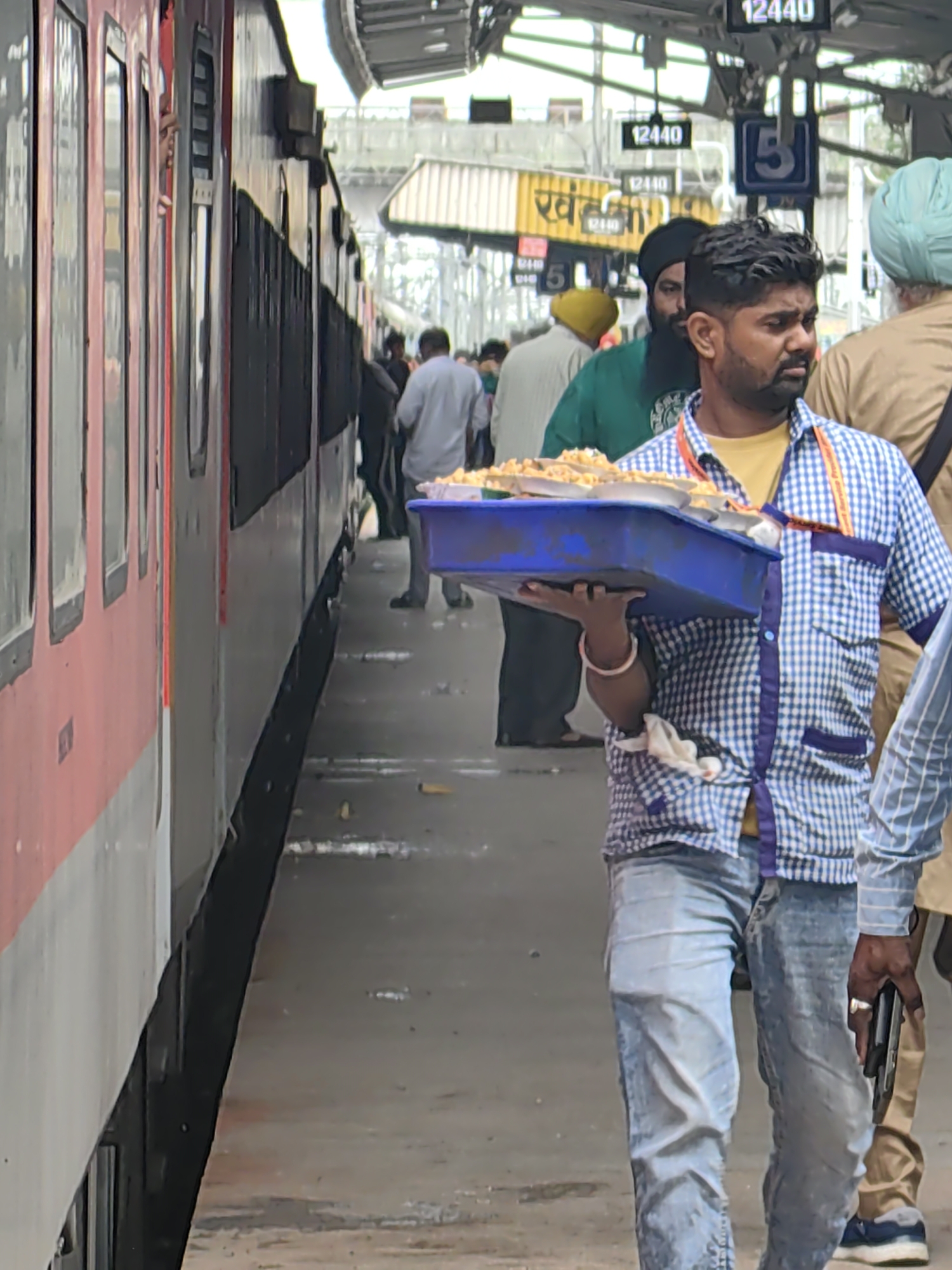 An Indian man in a blue and white checkered shirt and blue jeans walks along a train platform in Khandwa, India, holding a blue plastic tray filled with food. The train, red and white, is on the left side of the image. In the background, people are visible, and signs indicate the station name "Khandwa" and platform numbers. The platform is paved, and there are overhead structures and lighting. The man is looking to his right, and his left hand is on the tray, while his right hand is at his side. The overall scene depicts a busy railway station environment.