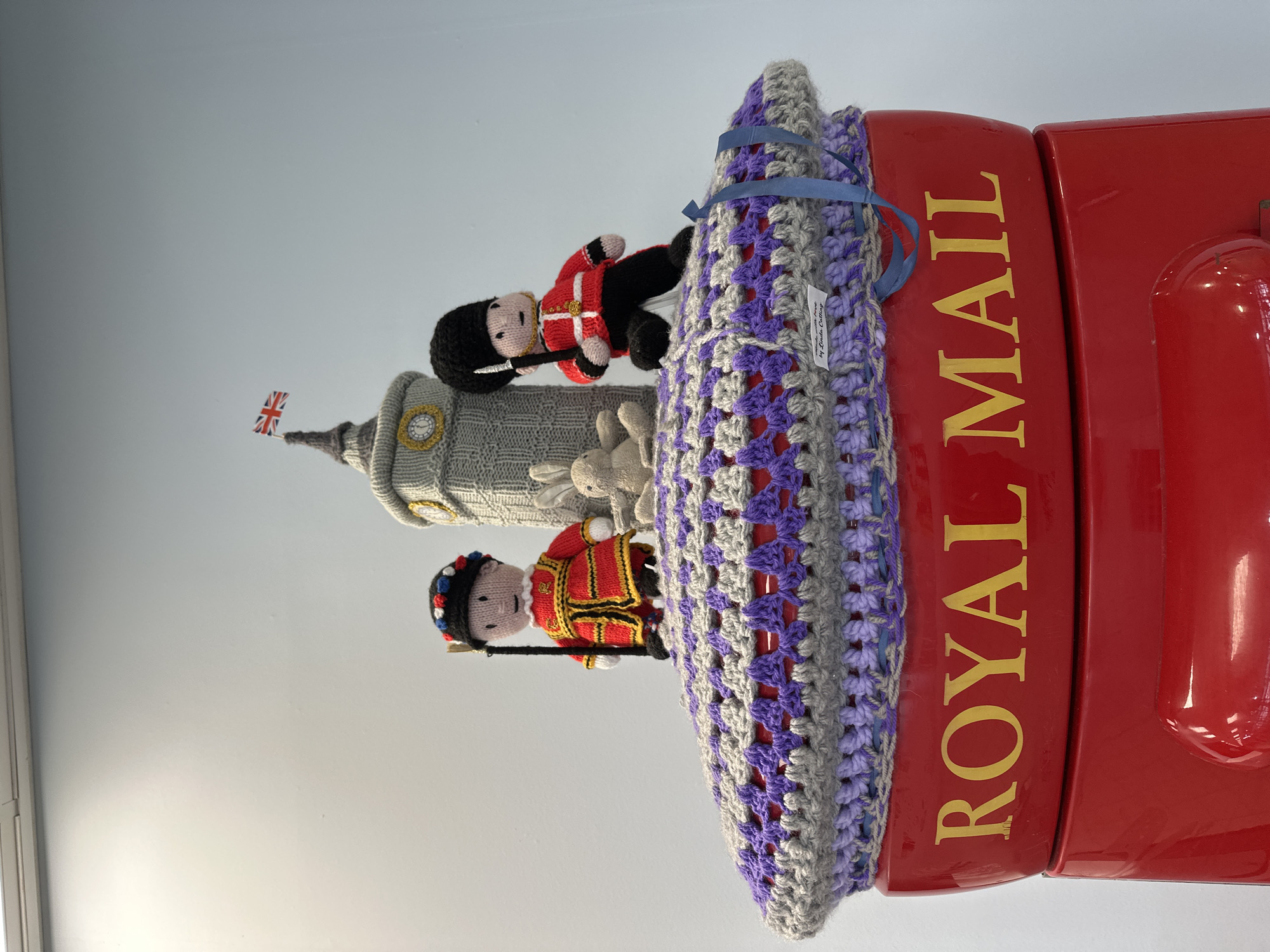 The image shows a crocheted topper on a red Royal Mail postbox. The topper features a depiction of Big Ben and two Grenadier Guard figures. The scene is likely located in Rochford, United Kingdom, given the UK postbox and London-themed topper. The topper is adorned with a blue ribbon. A small tag is visible on the topper.