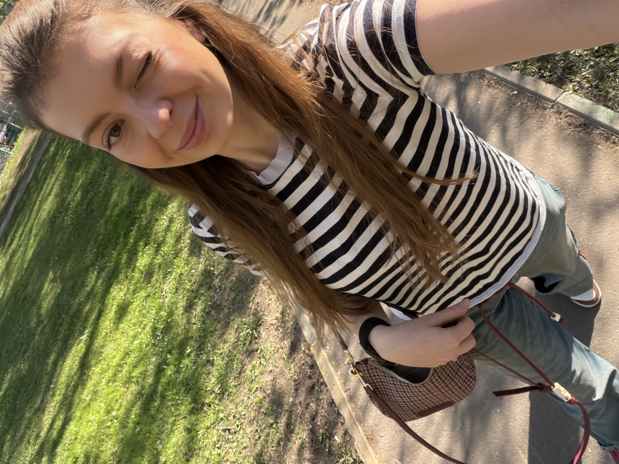 A young woman is standing outside on a sunny day. She is winking with her left eye closed. She is wearing a black and white striped shirt, jeans, a black watch on her left wrist and carrying a brown purse over her shoulder. The location appears to be a park with a grassy area and a sidewalk. It is possible that this is the city of Moscow, Russia.