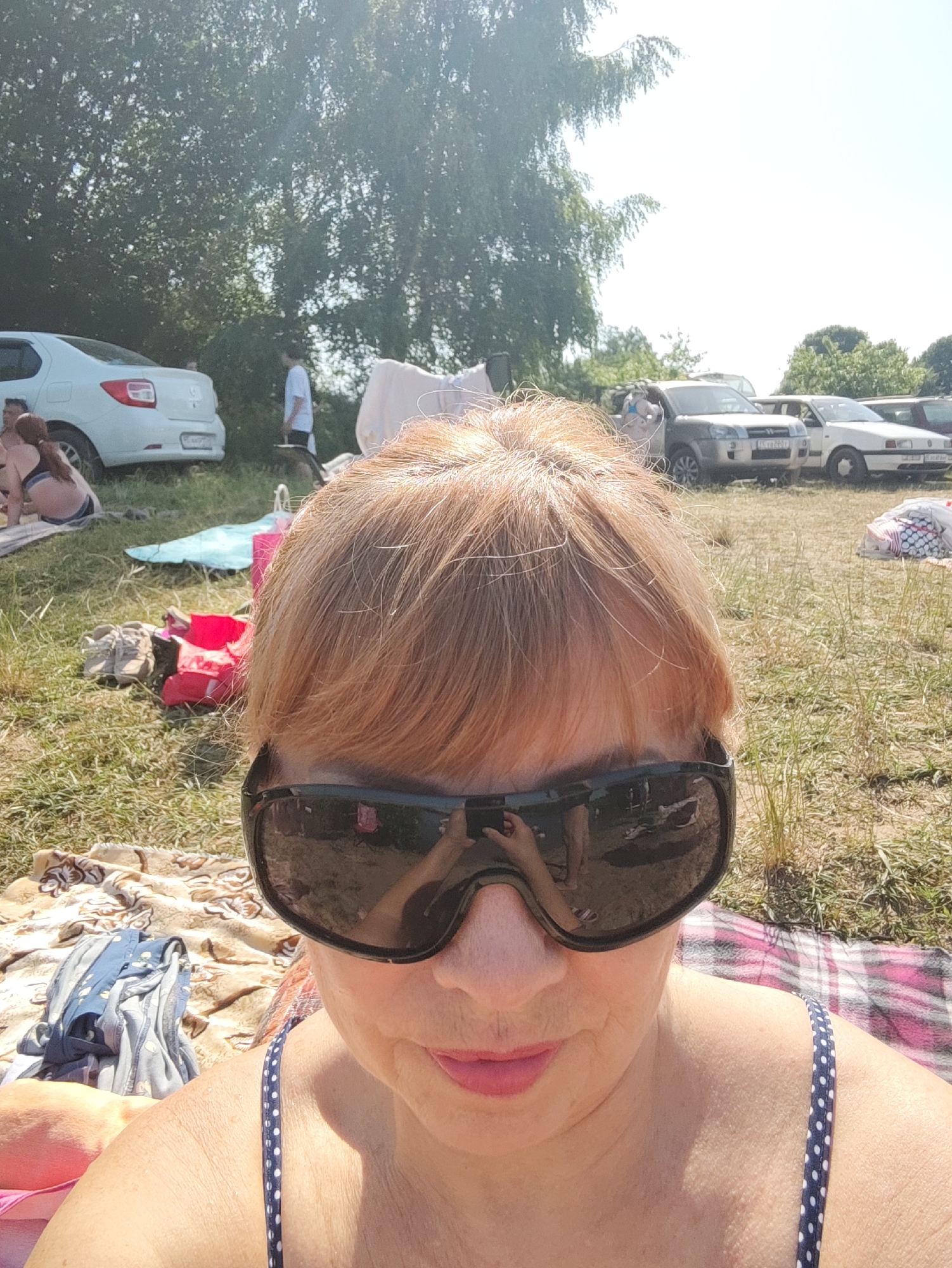 This is a selfie taken outdoors, likely during the daytime given the bright sunlight and shadows. The scene appears to be a gathering of people in a grassy, open area with trees in the background. Several cars are parked nearby, suggesting a picnic, outdoor event, or beach outing.
The foreground is dominated by a woman wearing large, dark sunglasses and a blue and white polka dot swimsuit. Her hair is a reddish-blonde color. She is looking towards the camera, with a slight smile on her lips. The sunglasses reflect the scene in front of her, showing a person holding a phone, likely the photographer.
In the middle ground, various objects are scattered on the grass, including towels, a blue mat, and some brightly colored bags, possibly picnic supplies. A few people are visible in the background, including a person in a white t-shirt and shorts walking away from the camera. Another person is seen sitting on the ground near a white car.
The overall atmosphere seems relaxed and summery. The weather appears to be warm and sunny, with clear skies. The location is identified as Rovnoe, Russia. No discernible text is visible in the image.