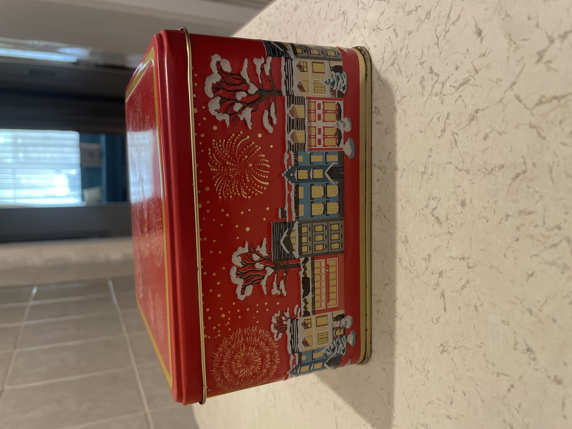 A rectangular red metal tin with a holiday-themed illustration sits on a speckled countertop. The illustration depicts a snowy winter village scene with buildings, trees covered in snow, snowmen, and fireworks in the sky. The tin has a gold trim along the top and bottom edges. The background shows a tiled wall and a doorway leading to an interior space with sheer curtains and furniture, suggesting a home environment. The scene is static and shows no people or activity.