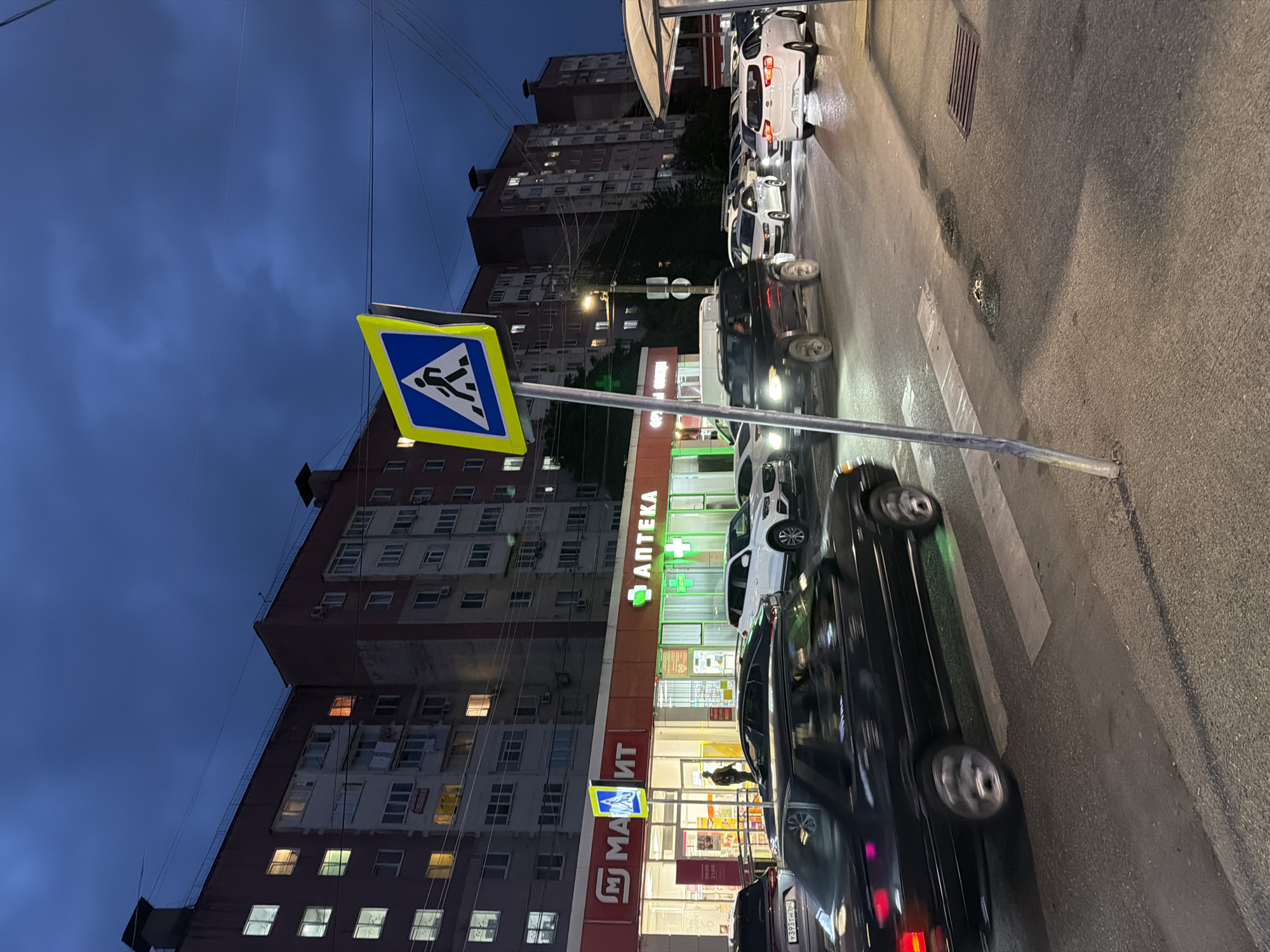 The image captures a dusk scene on a street in Sochi, Russia, likely in a residential or commercial area, given the presence of a multi-story apartment building and a storefront. A pedestrian crossing is prominent in the foreground, marked by white stripes on the asphalt. A bright yellow and blue pedestrian crossing sign stands to the left of the frame.
The street is busy with traffic, with several cars visible in motion and stopped. A black sedan is crossing the pedestrian area from the left, its headlights and taillights illuminated. To the right, a line of cars, including a white SUV and a white sedan, are stopped or moving slowly. The storefront appears to be a pharmacy, indicated by a prominent "АПТЕКА" (Apteka - Pharmacy) sign with a green cross logo. The building behind the storefront is a large, multi-story apartment complex with many windows, some of which are lit from within, suggesting occupancy. The sky is a deep blue, indicating twilight or evening, with some wispy clouds. Power lines and streetlights are also visible. The overall impression is of a typical urban scene during the transition from day to night.