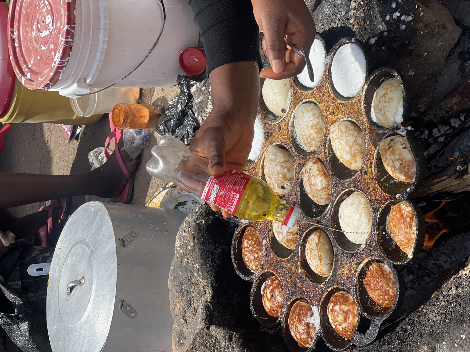 An overhead shot captures a person cooking small, round, pancake-like items outdoors in Jos, Nigeria, under bright midday sunlight. The individual, with dark skin, is wearing a black long-sleeved top and pink flip-flops. They are using a well-worn, multi-cavity cast-iron pan, similar to an aebleskiver pan, which sits directly over an open fire or hot coals.
The person's right hand holds a clear plastic bottle, from which yellow cooking oil is being poured onto the items in the pan. The left hand uses a metal utensil, possibly to turn or adjust the cooking items. The items in the pan are in various stages of preparation, ranging from white, uncooked batter to golden-brown, cooked surfaces.
The cooking setup is on a dirt or sandy ground. Surrounding the immediate cooking area are several basic containers: a white bucket with a red-splattered lid, other white and yellow buckets, and a large aluminum pot with a lid. Scattered nearby are plastic bags and other discarded items, typical of a street food vendor setup. Visible text on the oil bottle's label includes "GREAT TASTE" along with red and white printed nutritional information.