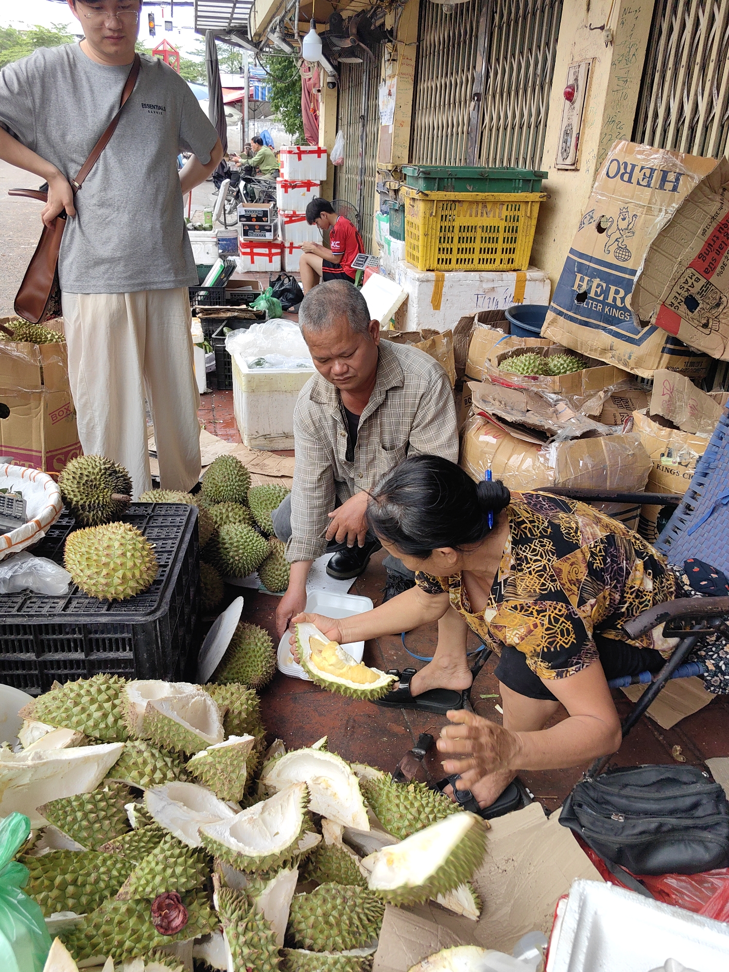 In a bustling outdoor market setting in Hà Nội, Vietnam, three individuals are engaged around a durian stall. The scene is brightly lit by daylight, suggesting daytime.
In the foreground, a woman wearing a patterned long-sleeved shirt and black shorts sits on a low chair, meticulously peeling a durian with her hands, revealing its pale yellow, pungent flesh. Beside her, a man in a plaid shirt squats, observing or assisting with the durian preparation, his hands near a white plastic plate. Numerous durians, both whole and partially opened, and a large pile of thorny green durian peels are scattered on the ground and in black plastic crates around them, indicating active sales and consumption.
Standing in the background, a young man wearing glasses, a grey "ESSENTIALS GARKIE" t-shirt, and light-colored pants observes the activity, with his hands on his hips. He carries a brown leather satchel over his shoulder.
The stall is set up against a yellow wall with a closed metal roller shutter, adorned with various markings including "H7 26" and "2726". Around the stall, cardboard boxes are stacked, some bearing the text "HERO FILTER KINGS" and others with partially visible Vietnamese text such as "Bảo quản sữa" (Store milk/Preserve milk), "Đậu trứng MASTER", and "khô th". One box clearly shows "BOX". Yellow and green plastic crates are also stacked against the wall. A handwritten "10.000/20$" is visible on one of the boxes, possibly indicating a price or quantity. The overall impression is one of a busy, functional street vendor operation.