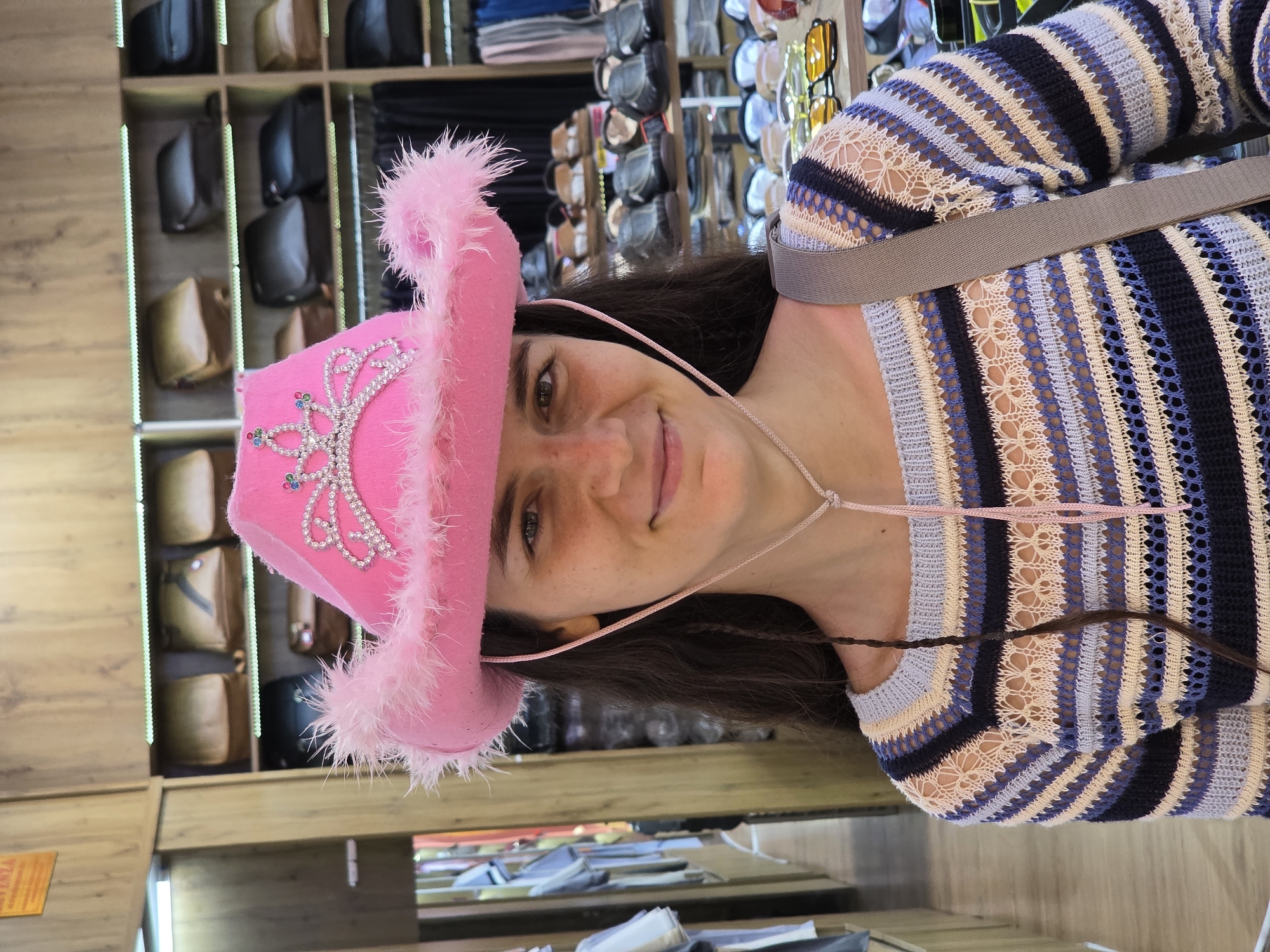 A woman with dark hair and a warm smile poses for a photograph indoors. She is wearing a bright pink cowboy hat, which features fluffy pink trim along the brim and a decorative, rhinestone-studded crown-like embellishment with small colored gems. A thin pink string from the hat is tied loosely under her chin. She is also wearing a long-sleeved, open-knit sweater with horizontal stripes in various shades of blue, white, and beige. A grey strap, possibly from a shoulder bag, crosses her chest.
The background of the image indicates an indoor retail setting, likely a shop or market stall in Phú Thủy, Vietnam. Tall, light-colored wooden shelves filled with numerous handbags or purses in various neutral colors such as black, brown, and tan are visible behind her. To the right, another display rack holds what appears to be sunglasses or eyewear. The woman looks directly at the camera, conveying a friendly and cheerful demeanor. A small, yellow sign with illegible text is partially visible in the bottom left corner of the frame. The image is oriented with the woman's head tilted to the right.