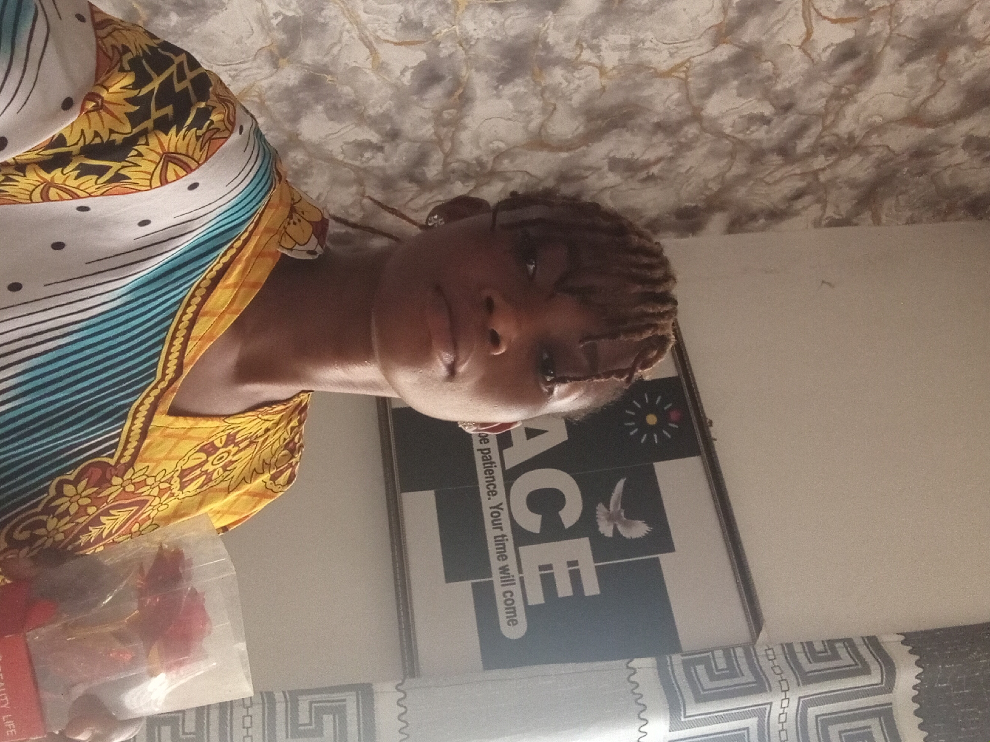 A person with braided hair is facing the camera. The person is wearing a patterned top with yellow, white, and blue stripes. A package of items is in the lower left corner. Behind the person is a wall with a framed print that says "ACE" and below that "Toe patience. Your time will come." The background is a white wall with a patterned ceiling. The location is Unknown, Unknown.