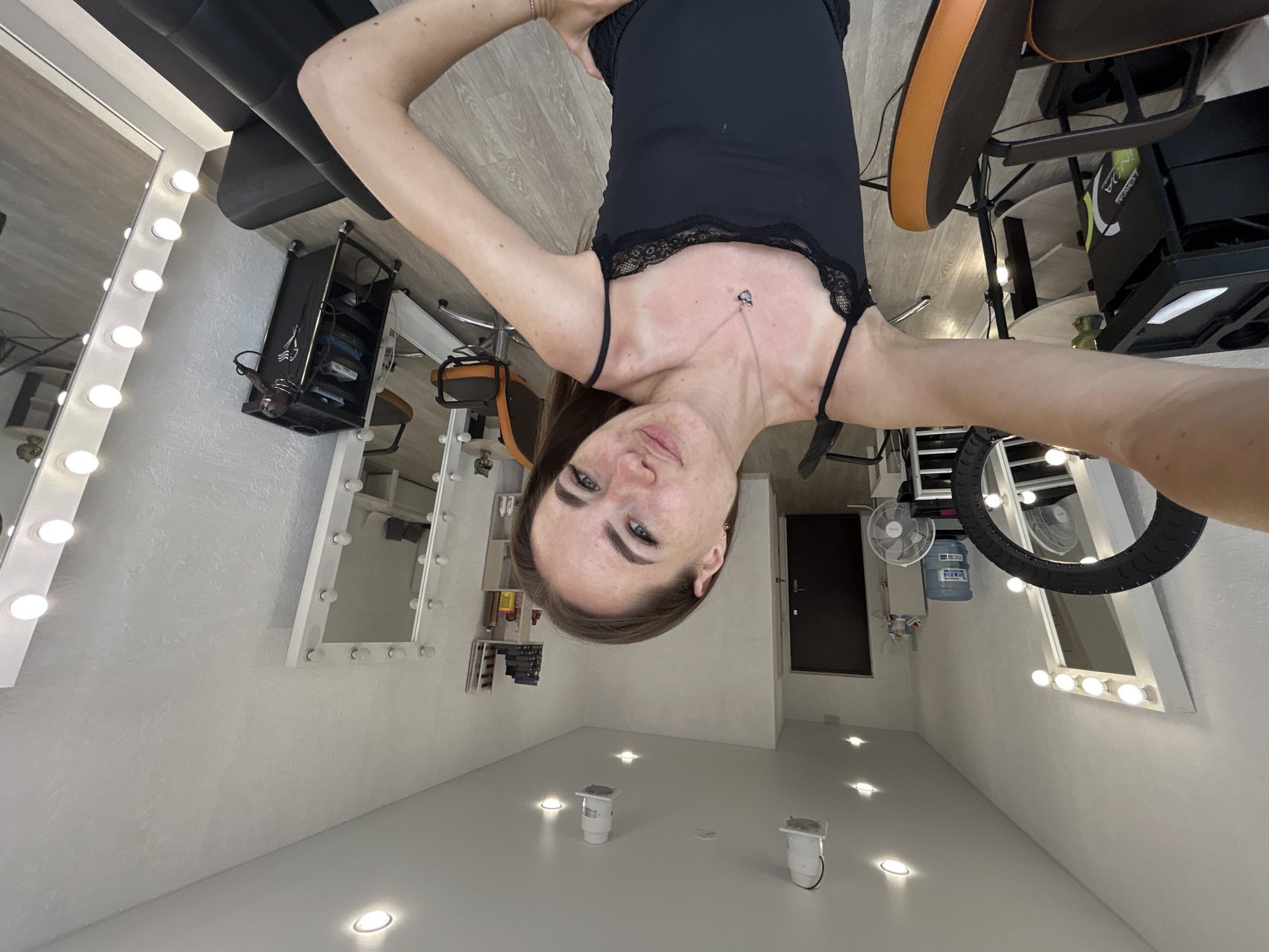 An indoor, eye-level, wide-angle selfie shows a young woman with medium brown hair and light skin, wearing a black tank top with lace trim and a silver necklace with a small pendant. She has freckles and appears to be posing for the photo. She is in what appears to be a beauty salon or makeup studio, indicated by several large vanity mirrors with bright lights, chairs, a hair dryer, and a multi-tiered cart. The room has white walls and a light-colored wooden floor. The woman's left arm is extended towards the camera, and her right arm is resting on her hip. The background shows several large mirrors, one with a ring light attached, and other salon equipment. There is a doorway visible in the distance.