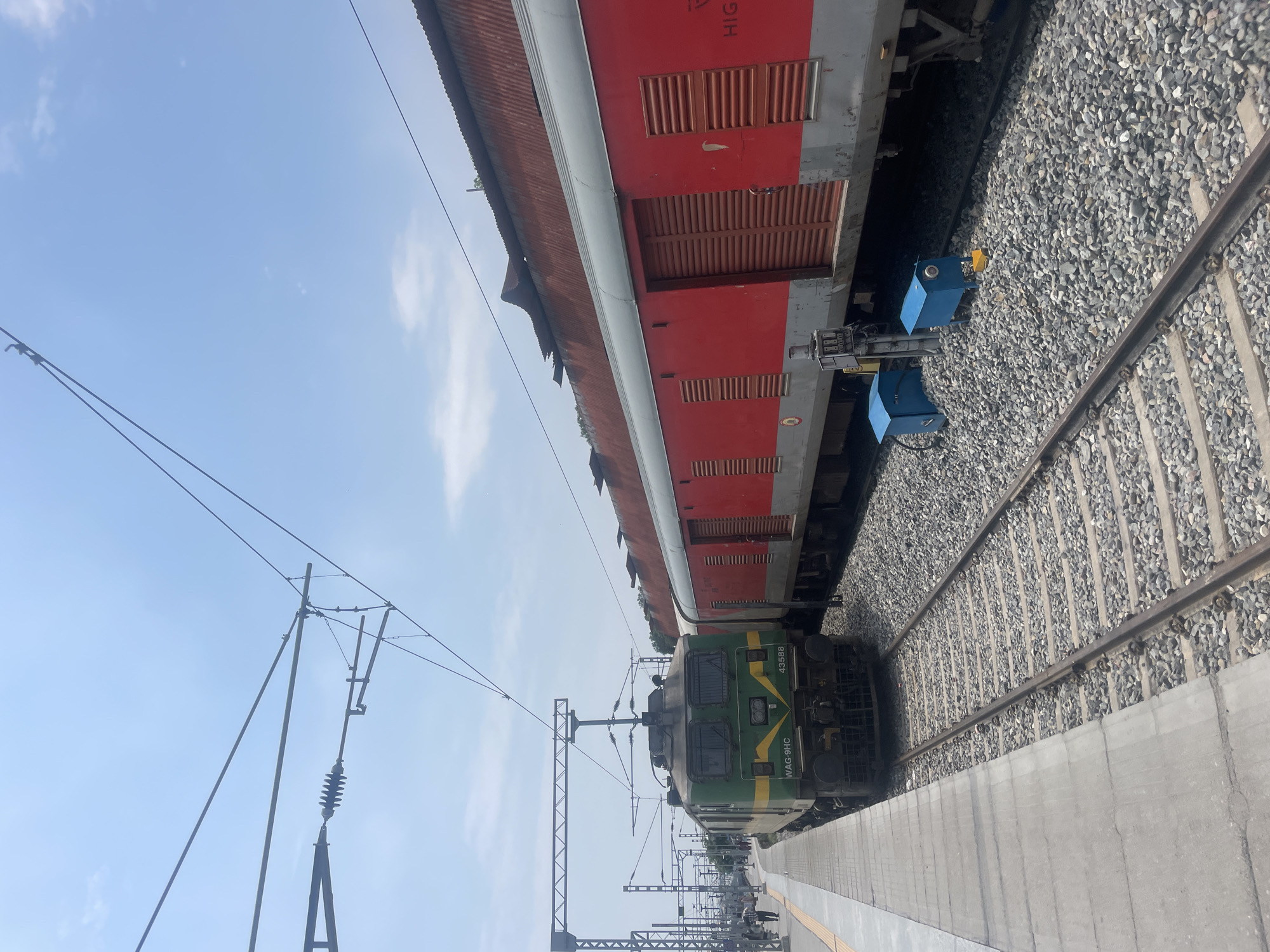 The image depicts a train station in Pathankot, India, under a clear blue sky with scattered clouds. A green and yellow locomotive, identified as WAG-9HC with the number 43588, is positioned on the left track. Adjacent to it on the right track is a red train car with a corrugated metal roof. The platform, made of concrete slabs, runs along the left side of the locomotive. The railway tracks are lined with gravel. Several electrical poles and overhead wires are visible, indicating the presence of an electrified railway system. Two small blue boxes with yellow elements are situated on the gravel between the tracks and the red train car. In the background, a few individuals can be seen walking on the platform, and other train cars are visible further down the tracks. The overall scene is that of a functional railway station during daytime.