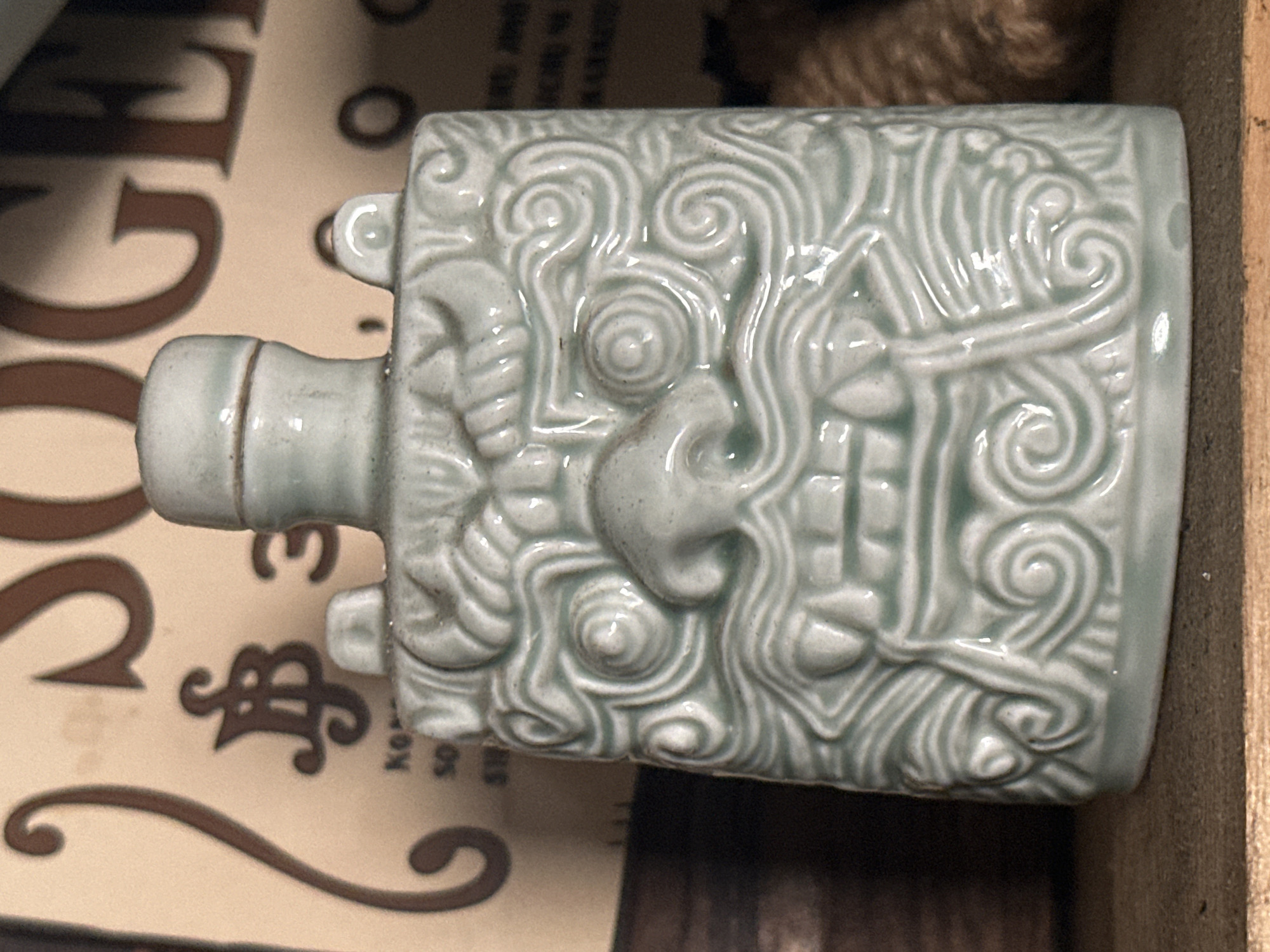 A pale green ceramic bottle, possibly a sake bottle, lies horizontally on a surface. It has a short, cylindrical neck with a ring and a square body decorated with raised, swirling patterns and a central, stylized face of an animal, possibly a dragon. The surface beneath the bottle is patterned paper, with large brown letters partially visible. The overall setting appears to be a shop or storage area, possibly in Andreyevka, Russia.