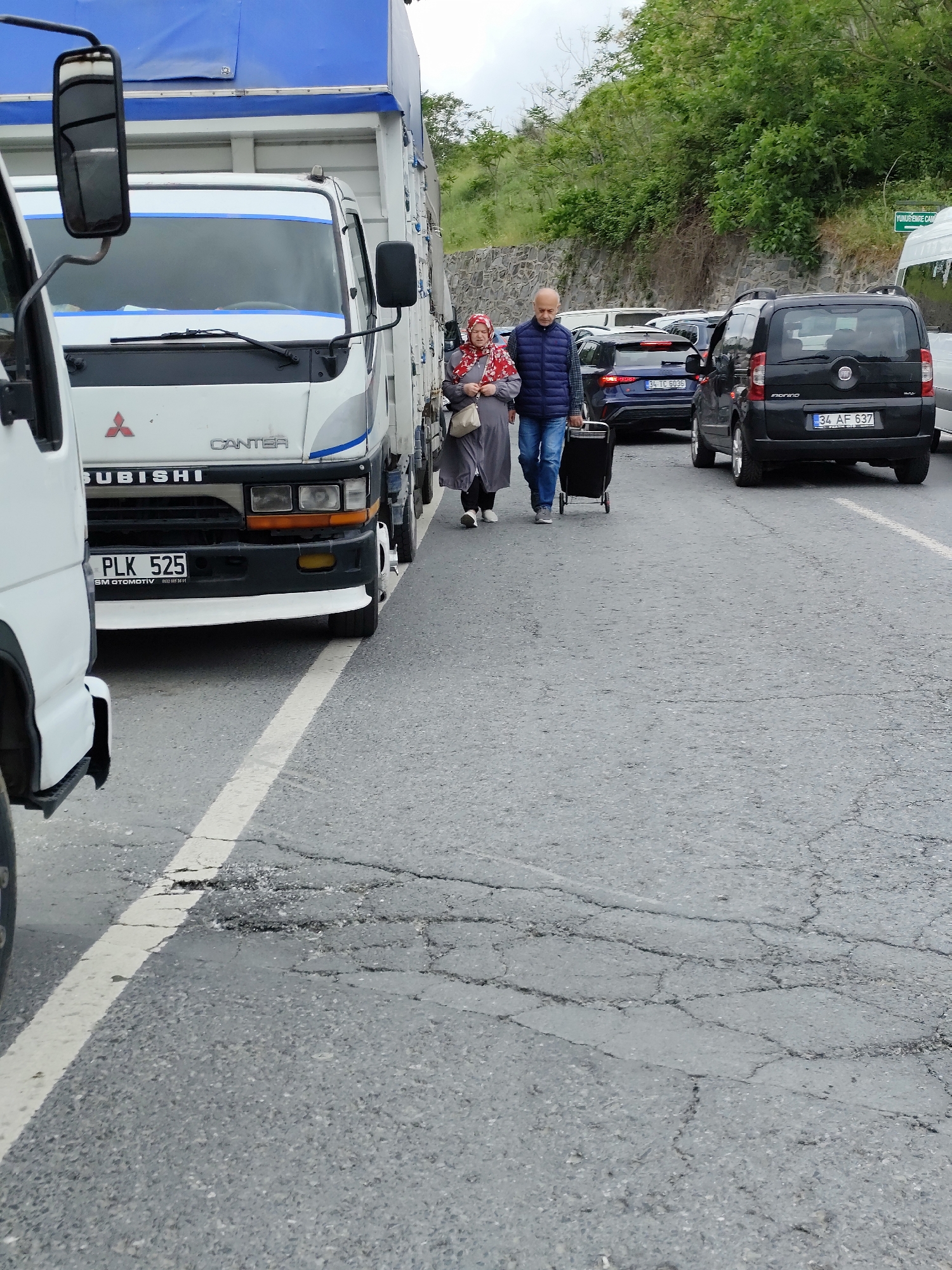 The image shows an outdoor urban scene on what appears to be a road shoulder or an area beside parked or stopped vehicles. A man and a woman are walking along the road's edge. The woman wears a headscarf and a long grey garment, carrying a purse. The man, wearing a blue vest over a collared shirt and jeans, pulls a small black rolling suitcase behind him. They are passing by a line of vehicles.
A Mitsubishi Canter truck is in the foreground, partially obscuring the view. In the background, there are several cars, including a black Fiat Qubo with license plate "34 AF 637" and a dark blue Audi with license plate "34 TC 6036". In the distance, there are green trees and foliage. A green road sign reading "YUNUS EMRE CAM" is partially visible. The road surface is grey and cracked. A white dividing line is visible along the road's edge.
