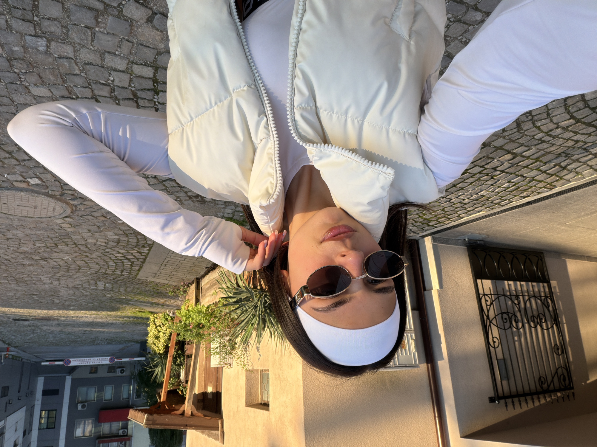 A selfie was taken by a young woman. She is wearing a white headband, sunglasses, a white long-sleeved shirt, and a white puffer vest. The background shows a building with barred windows, a house with a lush bush in front, and a modern building with a gate. The building with the gate is located in Gabrovo, Bulgaria.