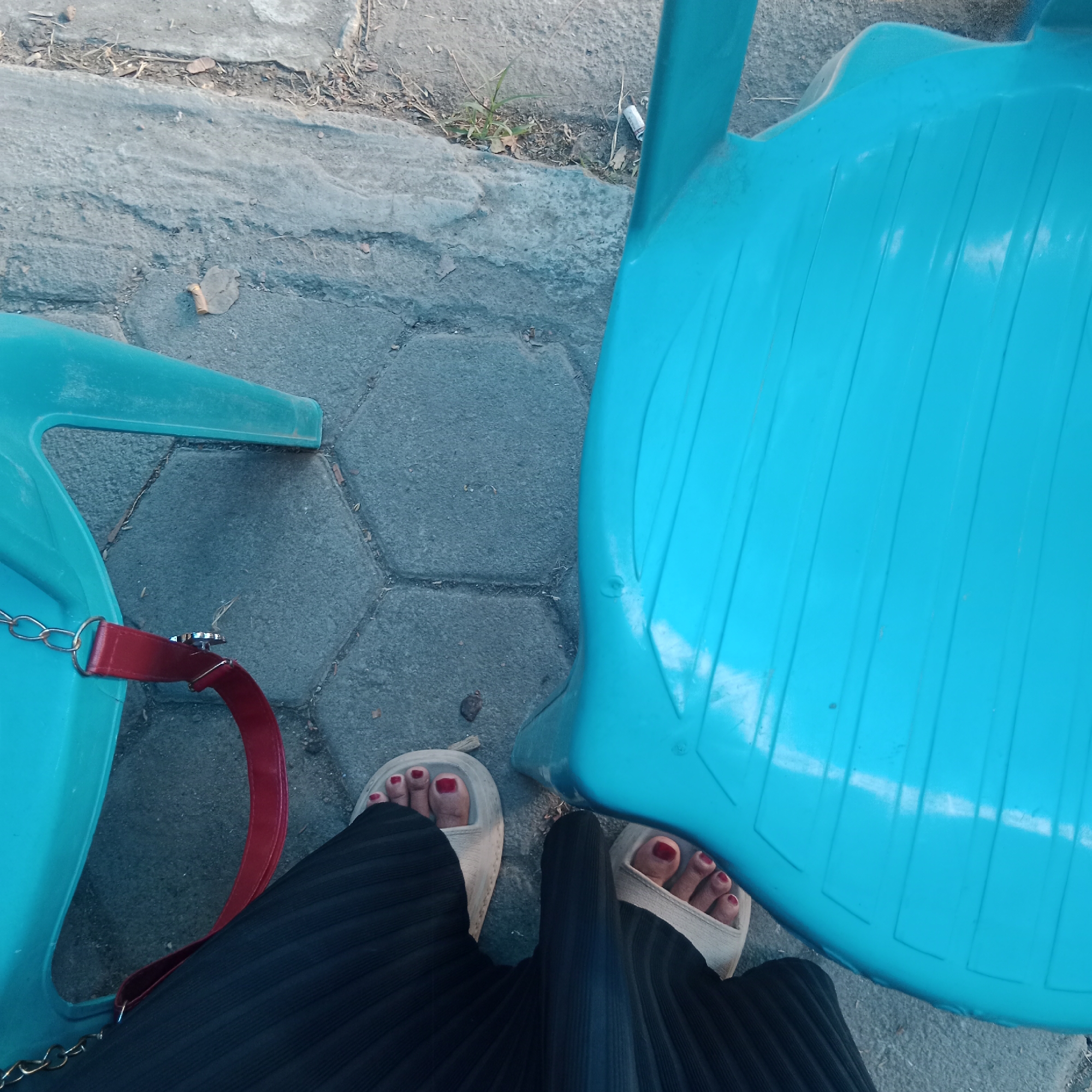 An overhead, medium shot captures a close-up view of a person's feet and legs, clad in a black pleated skirt and beige slide sandals, resting on a patterned concrete surface. The person is seated next to two bright blue plastic chairs, one of which is partially visible on the left, featuring a red strap attached to it with a metal clasp and chain. The other chair is more prominent on the right, its textured surface dominating the right side of the frame. The ground beneath is a mix of hexagonal and rectangular concrete pavers, with some debris, leaves, and a single cigarette butt visible between the paving stones. The scene is set outdoors, likely in a public area, given the presence of the plastic chairs and paving. The lighting suggests it is daytime, casting subtle shadows.