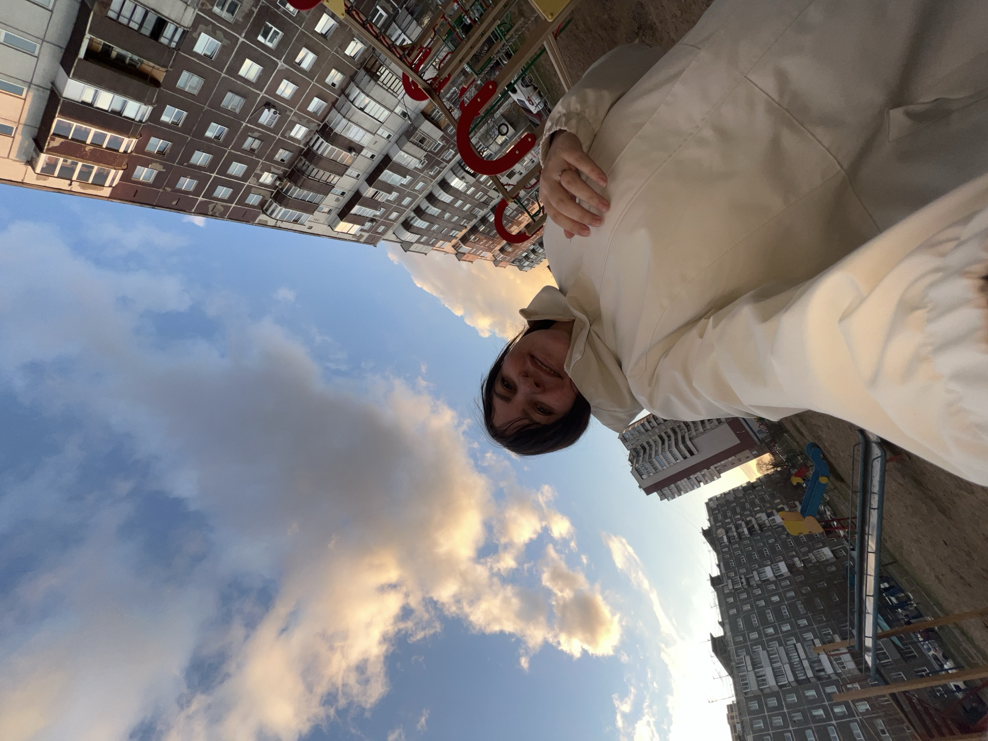 The image captures an upward-facing selfie taken in a residential area in Perm, Russia. The primary subject is a person, appearing to be a woman with dark hair and a light-colored (possibly off-white or beige) jacket, smiling faintly at the camera. Her hands are visible near her chest, one resting on the other.
The setting is an outdoor urban environment, likely a playground or courtyard surrounded by apartment buildings. In the background, several multi-story residential buildings with distinct, panel-like architecture, numerous windows, and small balconies are prominent. These buildings are characteristic of post-Soviet construction. Various playground equipment is visible, including red metal structures, a blue slide, and yellow elements suggesting a swing set or another play structure.
The sky occupies a large portion of the frame, showing patches of clear blue interspersed with scattered clouds. Some of these clouds are illuminated with warm, golden tones, indicative of late afternoon or early evening sunlight, possibly suggesting a sunset. The overall atmosphere is that of a typical residential district under partly cloudy skies. No visible text is present in the image.