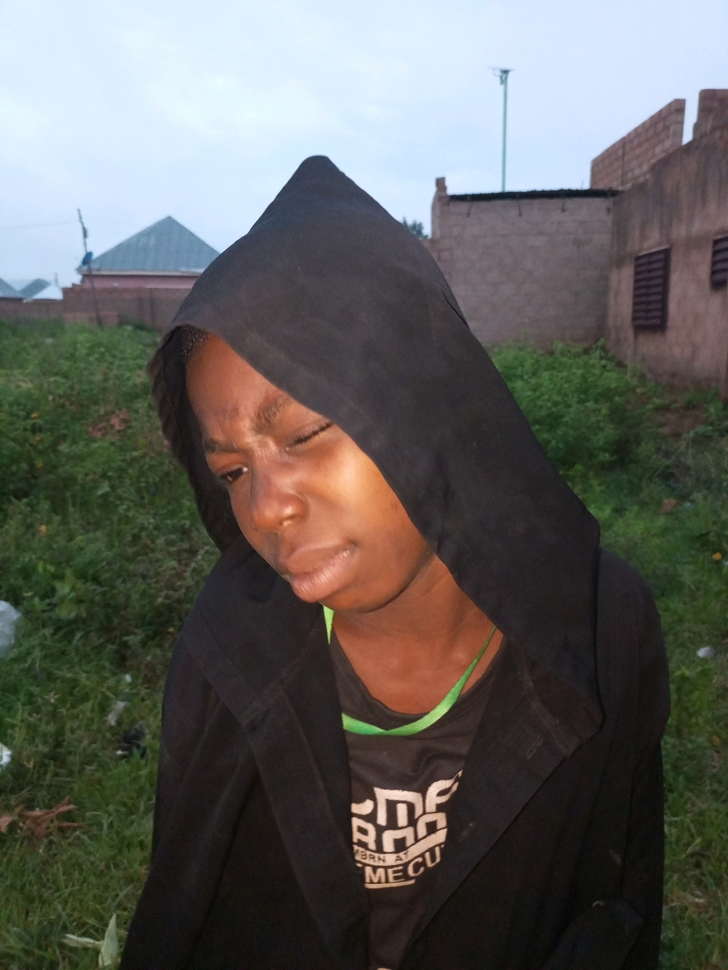 A person wearing a black hooded jacket and a black t-shirt with green trim is shown outdoors. The person's eyes are closed, and their expression suggests sadness or distress. The background includes grass, buildings with brick walls, and a light pole. The scene appears to be in an urban or semi-urban area.