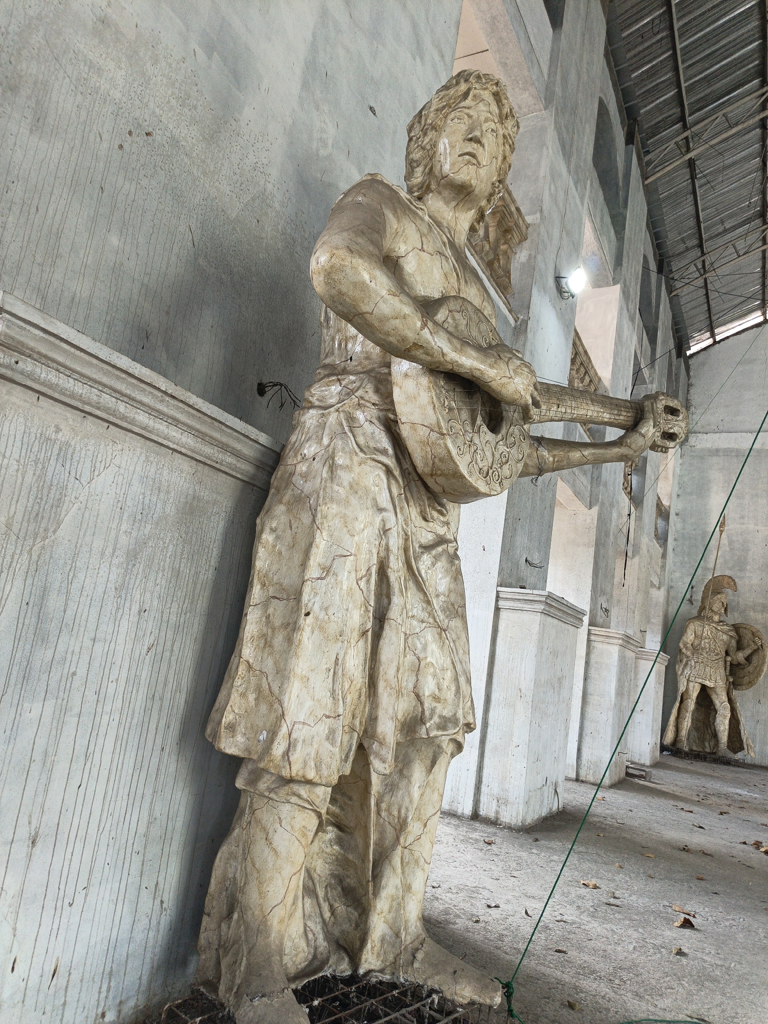 The image captures a statue of a man playing a lute, prominently displayed in what appears to be an outdoor covered structure. The statue, made of a weathered material resembling stone or plaster, exhibits numerous cracks and imperfections, suggesting age or exposure to the elements. The man is depicted with long, flowing hair, wearing a draped garment that falls to his knees. His hands are positioned as if playing the lute, which is intricately decorated.
In the background, another statue is visible, this one of a soldier in armor holding a shield and spear. This soldier statue is smaller than the lute player and stands further back in the frame, partially obscured by columns. The environment suggests an open-air pavilion or an unfinished architectural space, with a corrugated metal roof supported by visible beams and columns. The walls are a light gray, with signs of water staining and streaks running down them, indicative of an outdoor or semi-exposed location. The ground is concrete, scattered with dry leaves. A green rope is visible in the foreground, extending from the bottom left corner towards the right side of the frame, secured to the floor with a knot. The overall impression is one of an art installation or a public space featuring classical statuary, possibly in a state of disrepair or undergoing renovation. The specific location in Cibogo, Indonesia, suggests a regional cultural context for these sculptures.