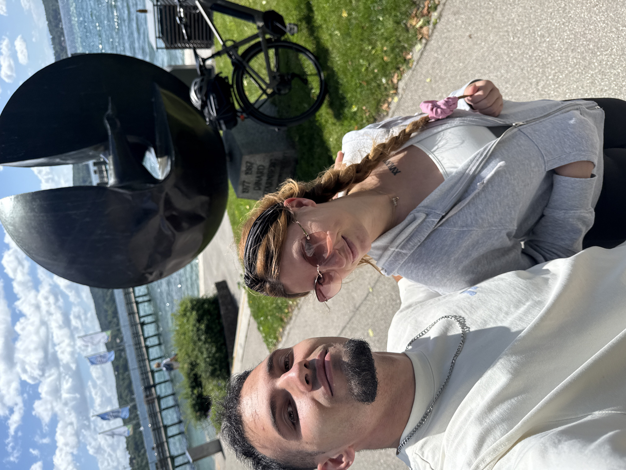 A man and a woman, appearing to be a couple, are taking a selfie outdoors on a sunny day. They are standing on a paved walkway next to a grassy area. In the background, a large abstract sculpture made of dark material is visible. To the right of the sculpture, a bicycle is parked. Beyond the sculpture and bicycle, a body of water, likely a lake, is present, with a wooden pier extending into it. Several flags are flying on poles along the pier. In the distance, hills covered in trees can be seen. The man in the foreground is wearing a light-colored t-shirt and a silver chain necklace. He has short dark hair and a goatee. The woman next to him has long braided blonde hair and is wearing a grey hoodie over a white top and sunglasses. She is holding a pink scrunchie in her hand. The overall scene suggests a relaxed outing at a scenic location, possibly a park or promenade along the Starnberger See in Germany.