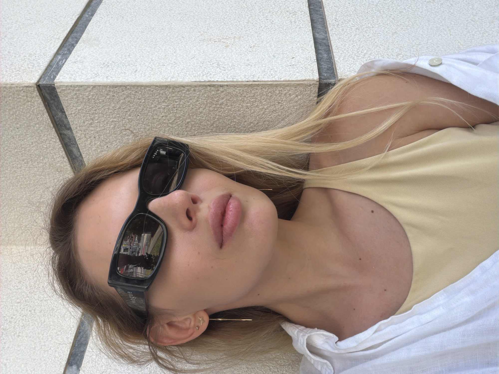The image is a close-up portrait of a blonde woman wearing black Vogue sunglasses, a light yellow tank top, and an open white linen shirt. She is leaning against a textured light-colored wall with dark horizontal trim, suggesting an outdoor urban setting. A reflection in her sunglasses shows a street scene with buildings and trees. She has a delicate gold earring with a star charm and a thin chain with a bar. The lighting suggests it is daytime, possibly outdoors in a city like Budva, Montenegro, given the architecture.