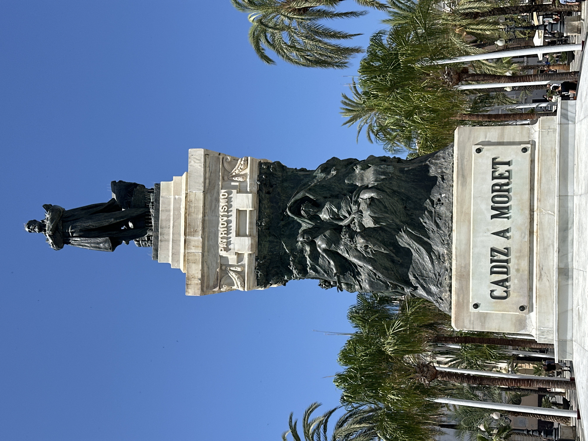 The image shows the Monument to the Heroes of the Constitution of 1812 in Plaza de España, Cádiz, Spain. The monument features a large bronze statue of a man on a pedestal, adorned with allegorical figures and inscriptions. "PATRIOTISMO" is visible on one side of the pedestal, and "CADIZ A MORET" is inscribed on the base. Tall palm trees line the square, and the sky is a clear blue. A few people are visible in the background, walking or standing in the plaza. The monument is made of marble and bronze, and it is a significant landmark in Cádiz.