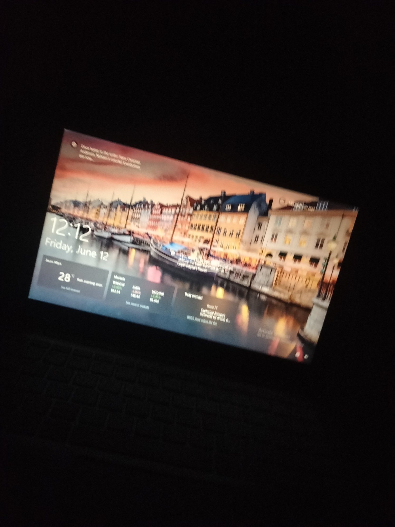 A laptop screen displays a lock screen with a picturesque canal scene featuring buildings and boats. The time is 12:12 on Friday, June 12. Weather information indicates 28 degrees Celsius with rain starting soon. Market data for WINDOW, AWDL, and USD/INR is visible. A section titled "Daily Wander" suggests content about "Dunes IV: Capturing Europe's Waterfalls by drone." The scene is dimly lit, and the laptop appears to be placed on a dark surface, possibly a keyboard.