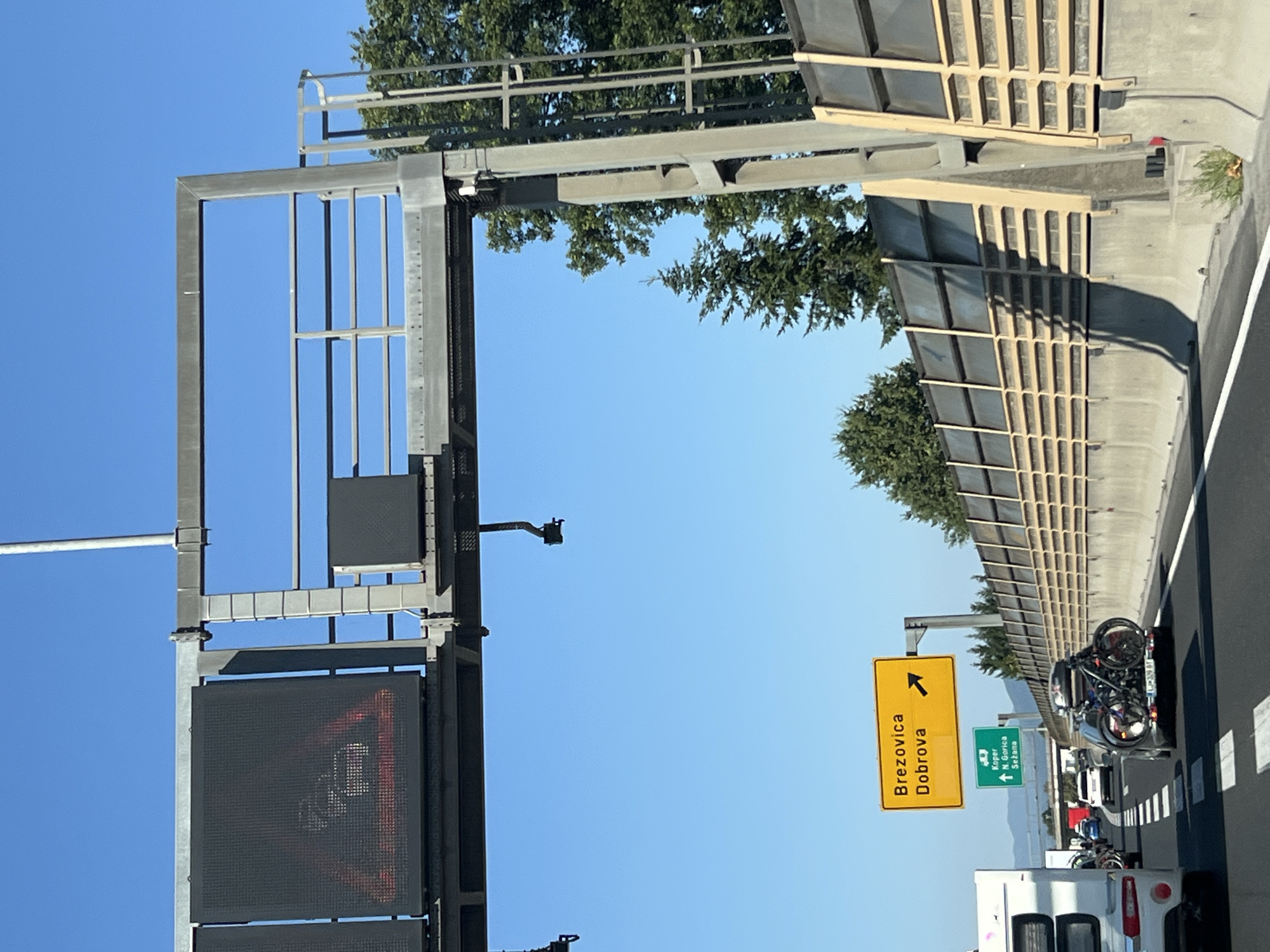 The image captures a scene from a highway, likely in Žeje, Slovenia, based on the road signs. The perspective is from inside a vehicle, looking forward. The sky is a clear, bright blue, indicating a sunny day.

Dominating the upper portion of the frame is a large overhead gantry structure, supporting electronic traffic signs and various pieces of equipment. On the left, a digital traffic sign displays a warning symbol, appearing to show a car with red flames, possibly indicating a hazard or traffic congestion. To its right, another black rectangular panel is mounted on the gantry. Hanging from the gantry are two cameras, one on each side, positioned to monitor the road below.

Below the gantry, a yellow directional sign points to the right, indicating "Brezovica" and "Dobrova". Further down and slightly to the left, a green sign shows an upward arrow and lists destinations: "Koper", "N. Gorica", and "Sežana".

The highway itself has multiple lanes, with white lane markings visible. Several vehicles are on the road, including a white camper van on the left, a dark-colored car with two bicycles mounted on a rack at the rear in the right lane, and other cars further ahead. A concrete sound barrier lined with wooden elements runs along the right side of the highway. Green trees and foliage are visible beyond the sound barrier and in the distance. The road surface appears to be asphalt.